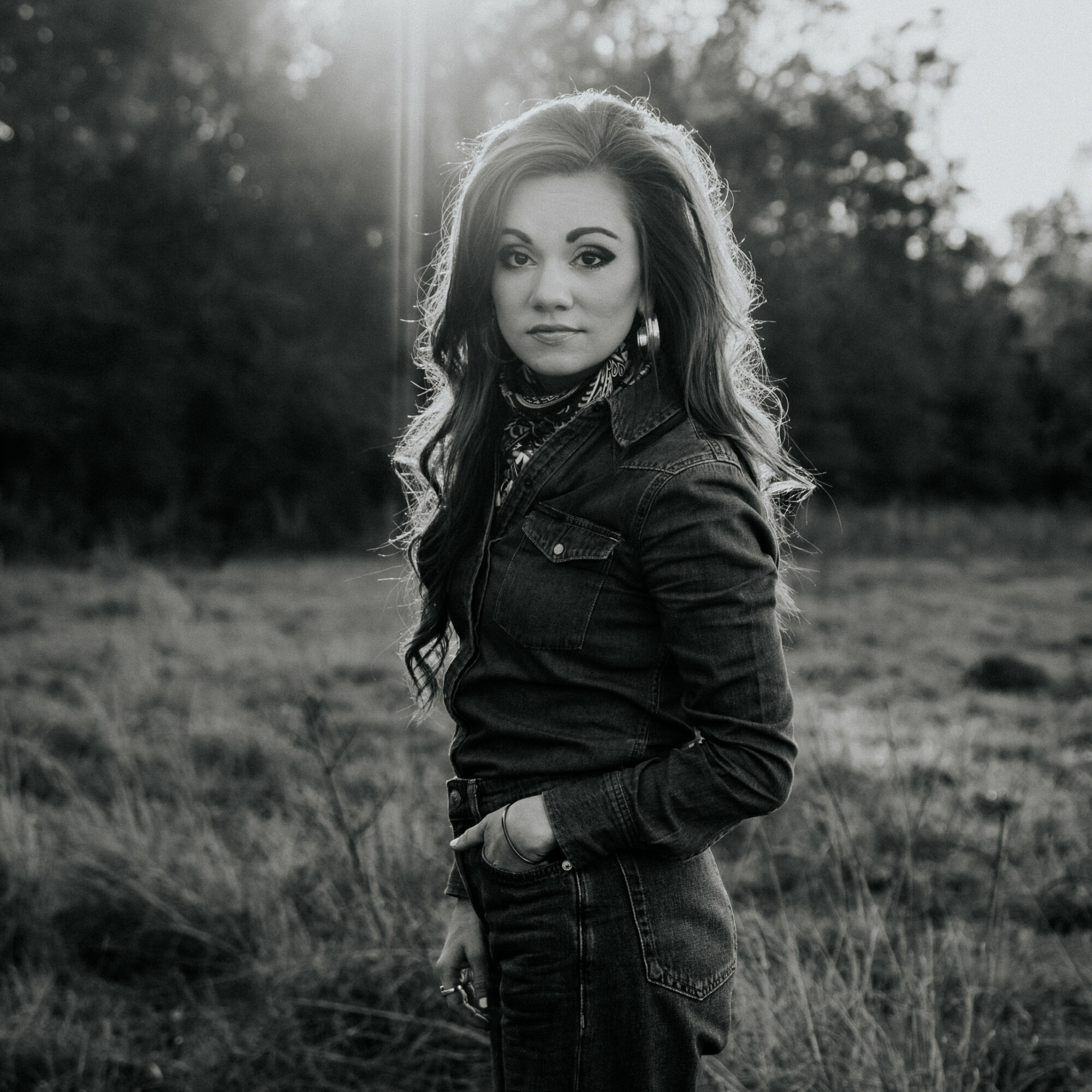 Woman with long hair standing outdoors in a field, wearing a dark jacket, with sunlight behind her, in black and white.