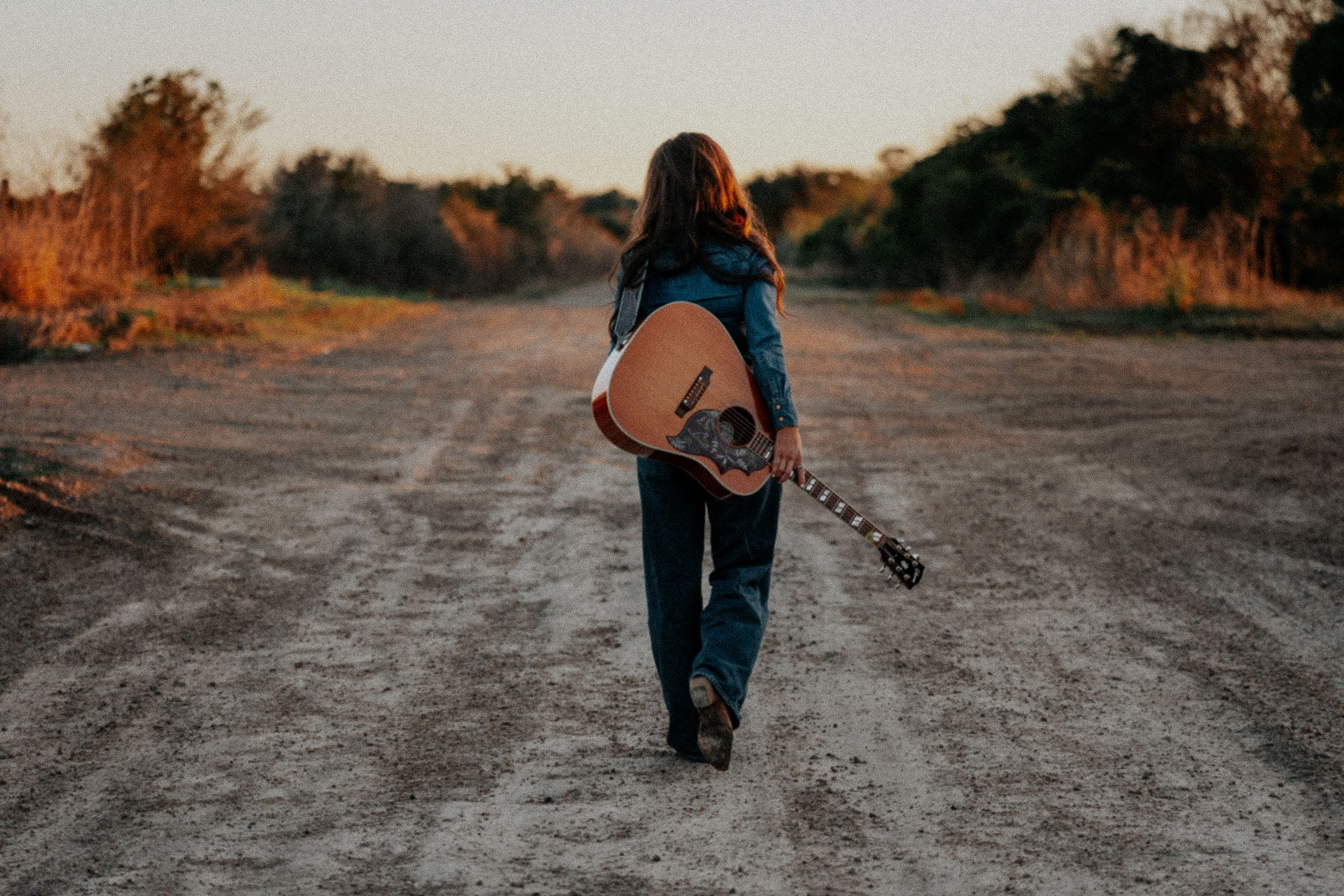 Person walking on a dirt road holding an acoustic guitar, with trees and sky in the background.