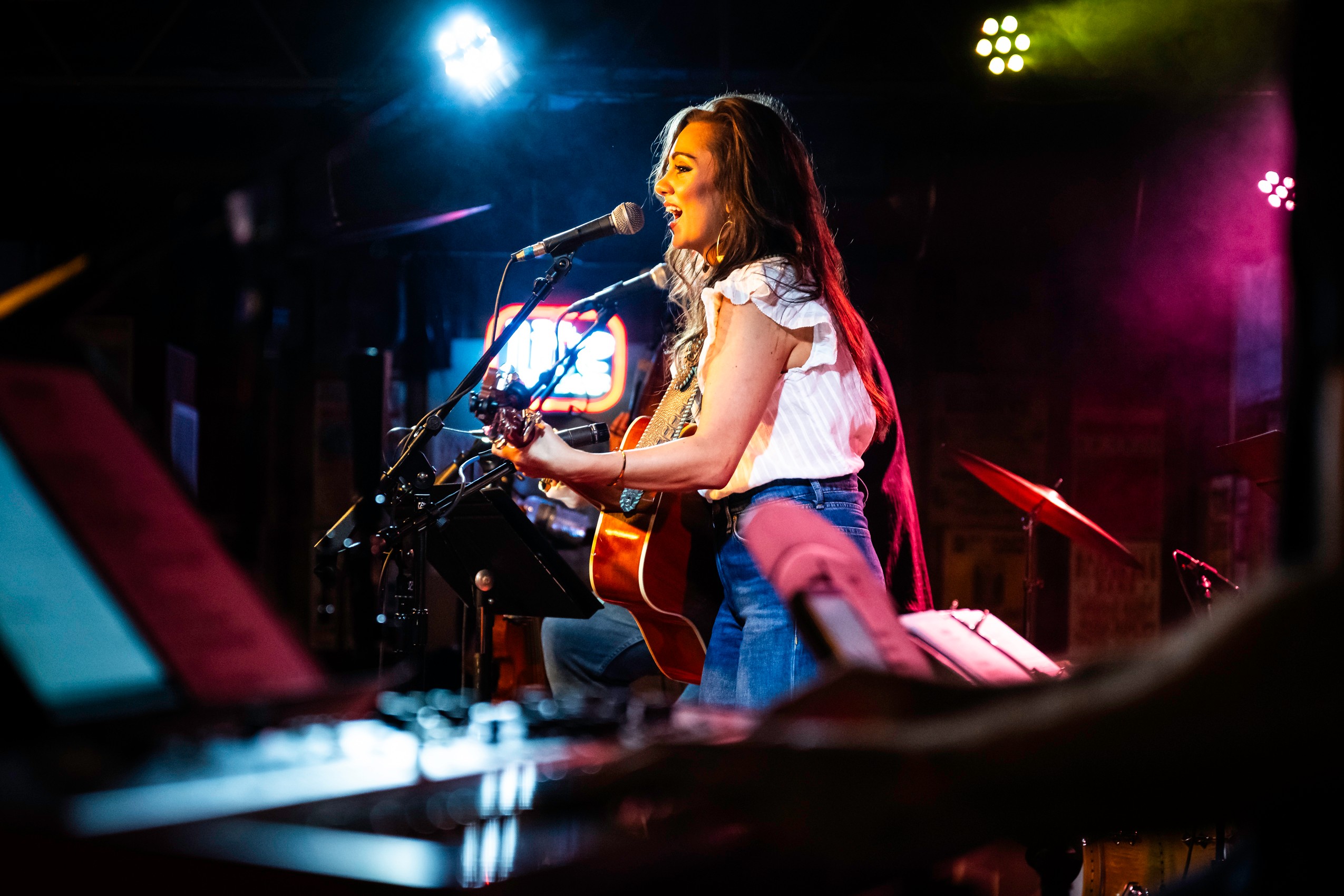 Young woman singing and playing guitar on stage with colorful lights, microphone, and music equipment visible.