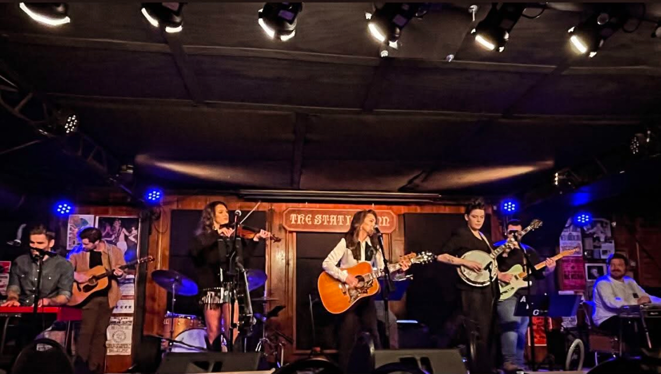 Group of six musicians performing on stage with guitars and microphones, stage lit with blue and purple lights, wooden backdrop.