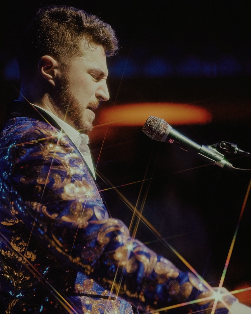 Man with curly hair and beard speaking into a microphone on stage, wearing a colorful jacket, with a dark background.