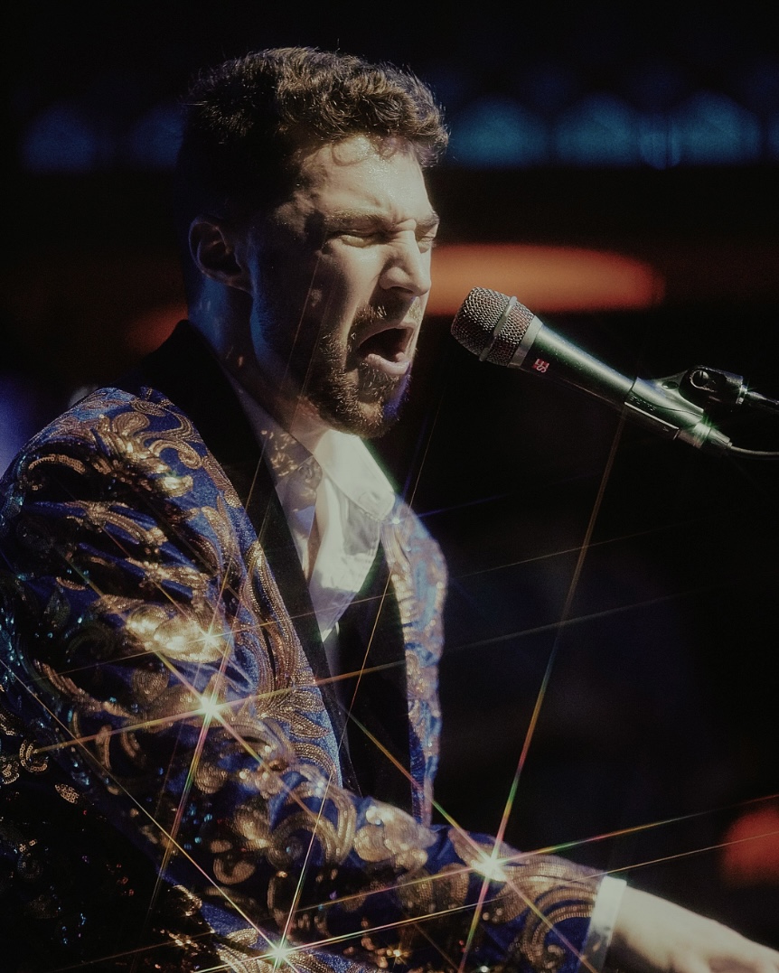 Man singing passionately into a microphone, wearing a shiny, patterned jacket, with a dark background.