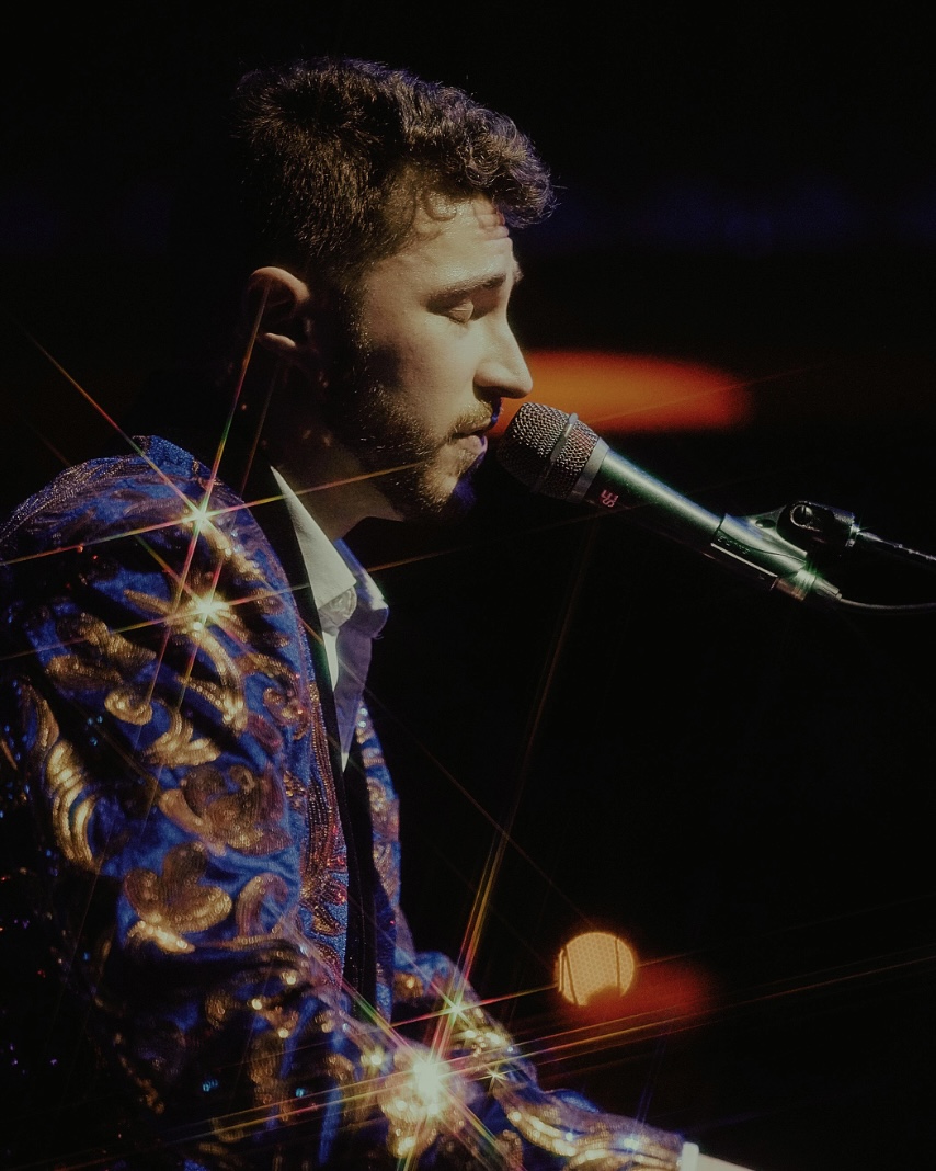 Young man singing into a microphone with a dark background, wearing a patterned jacket, illuminated by stage lights.