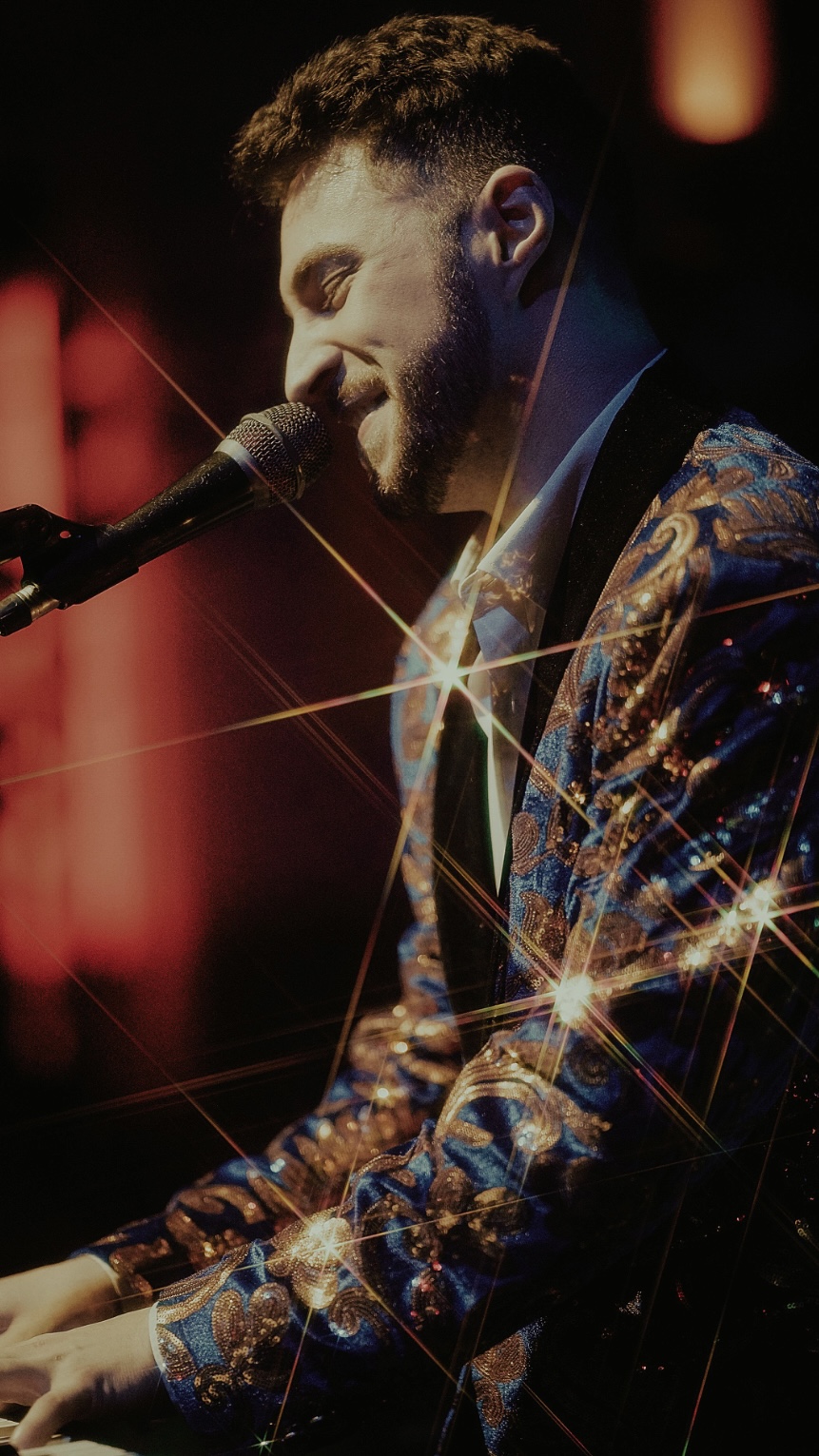 Male singer with a beard performs at a microphone, wearing a patterned jacket, with stage lighting effects.