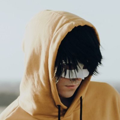 Person wearing a beige hoodie with the hood up, face partially obscured by dark hair and a white eye mask, neutral background.