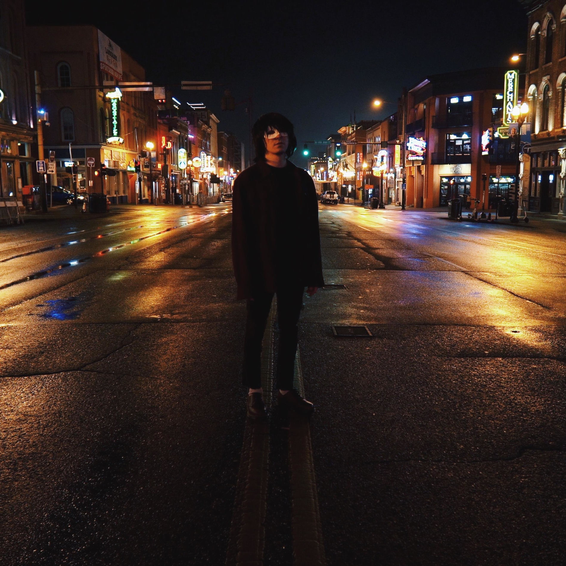 Person standing in the middle of a wet city street at night, illuminated by colorful neon lights on buildings.