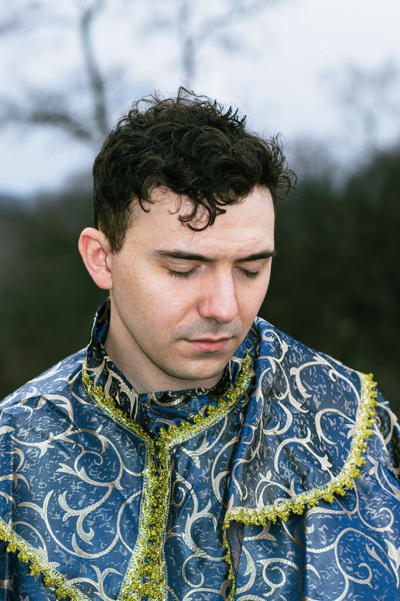 Young man with curly hair wearing a patterned blue and gold costume outdoors, looking downward.