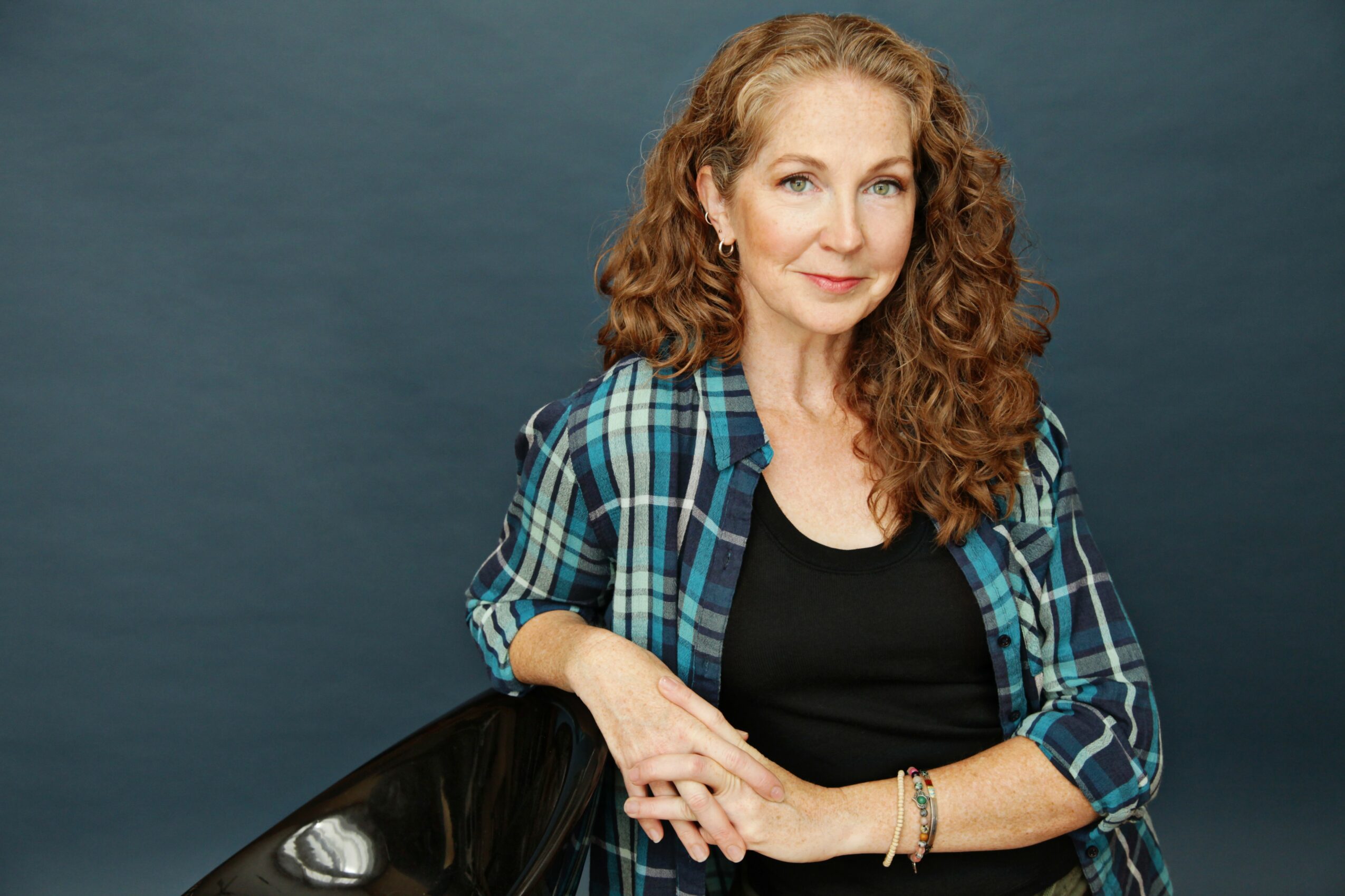Woman with curly hair wearing a plaid shirt and black top, smiling, against a dark background.
