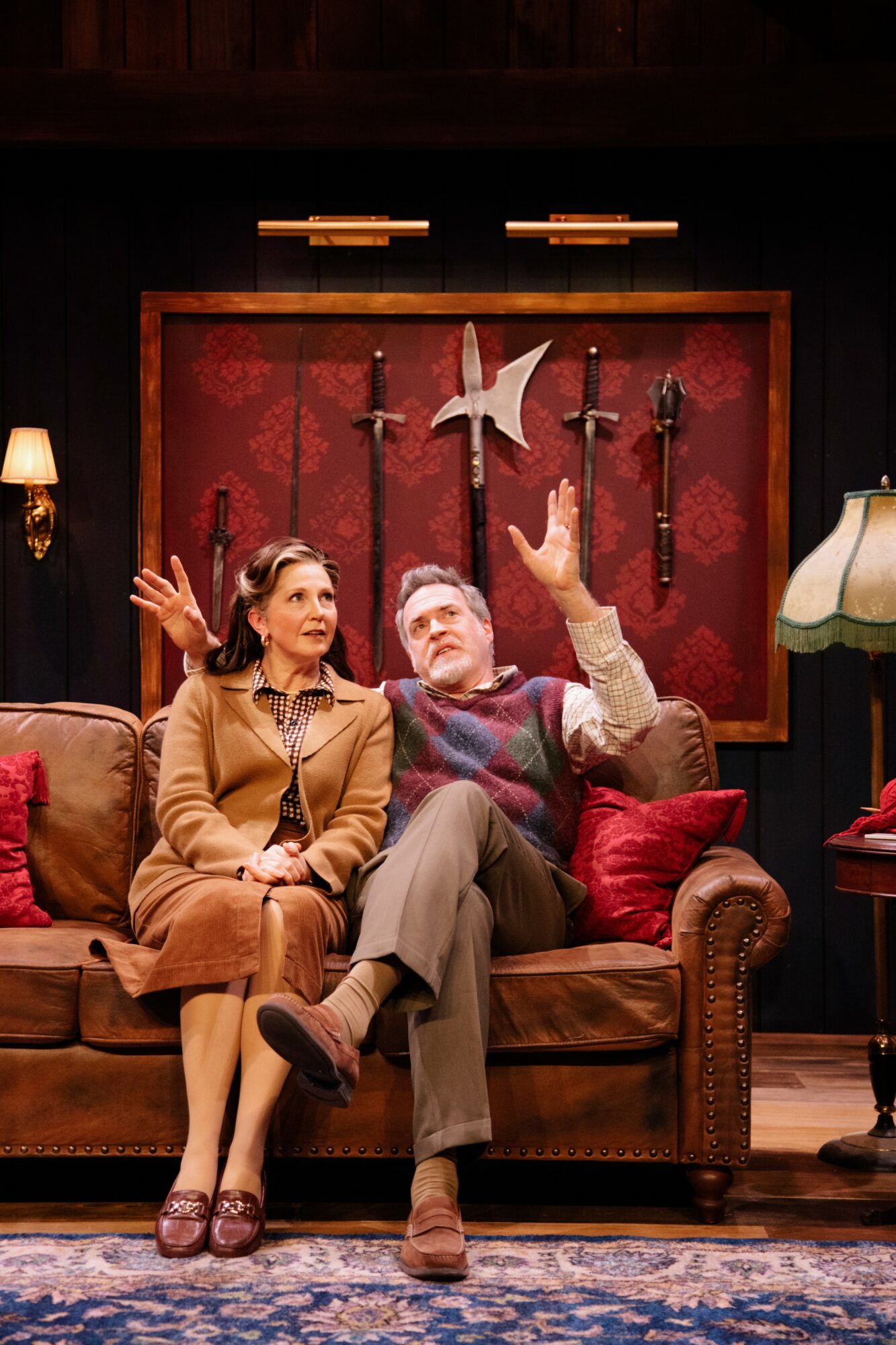 Two people sit on a sofa in a room with dark walls, red accents, and decorative swords on the wall behind them.