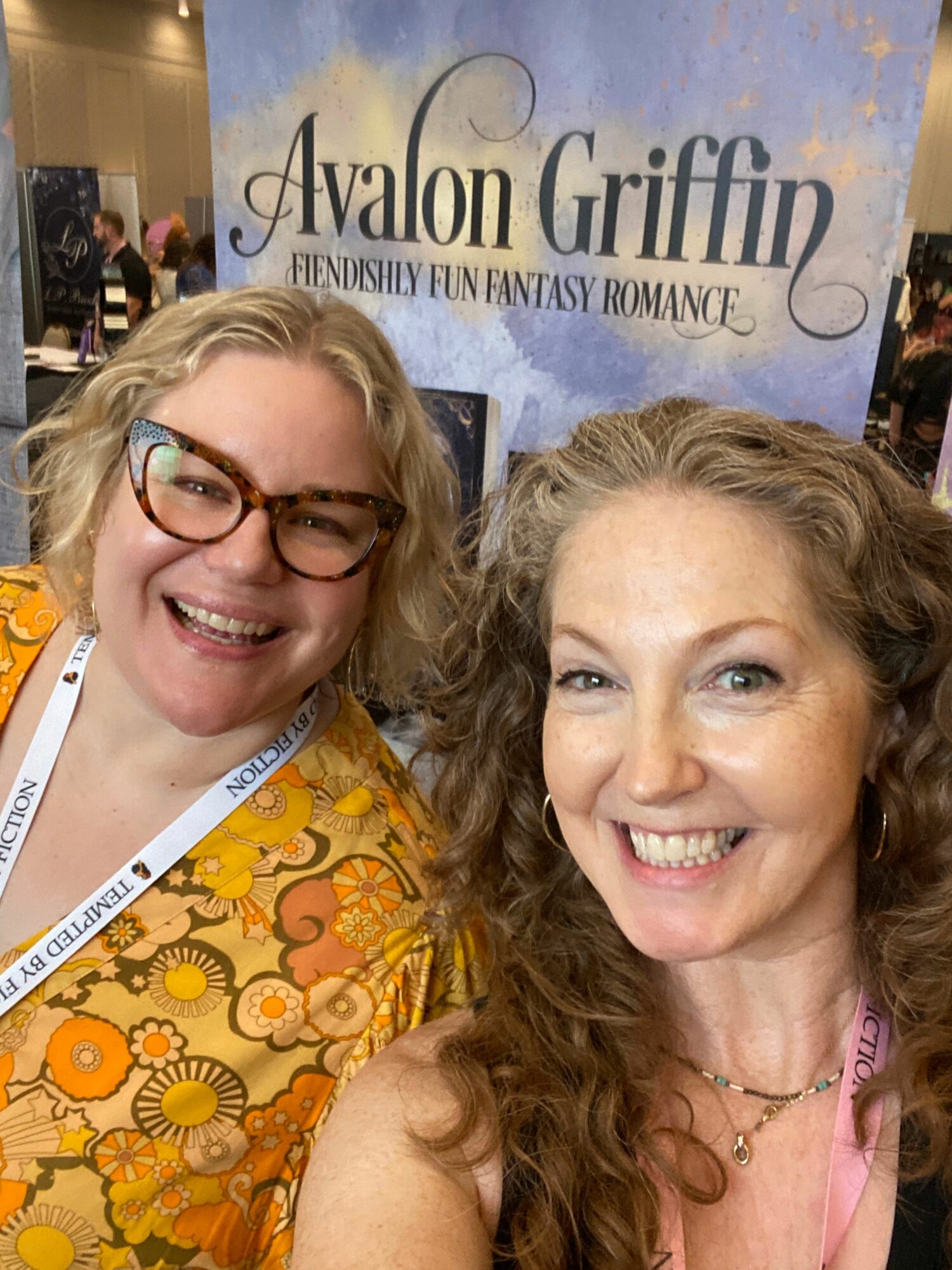 Two women smiling, one wearing glasses and a floral dress, the other with curly hair and a necklace, in a close-up shot.