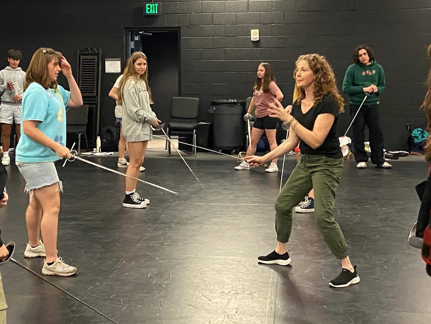 Group of people in a dance studio, some holding ropes, with a woman leading a dance move, black walls, and chairs in background.
