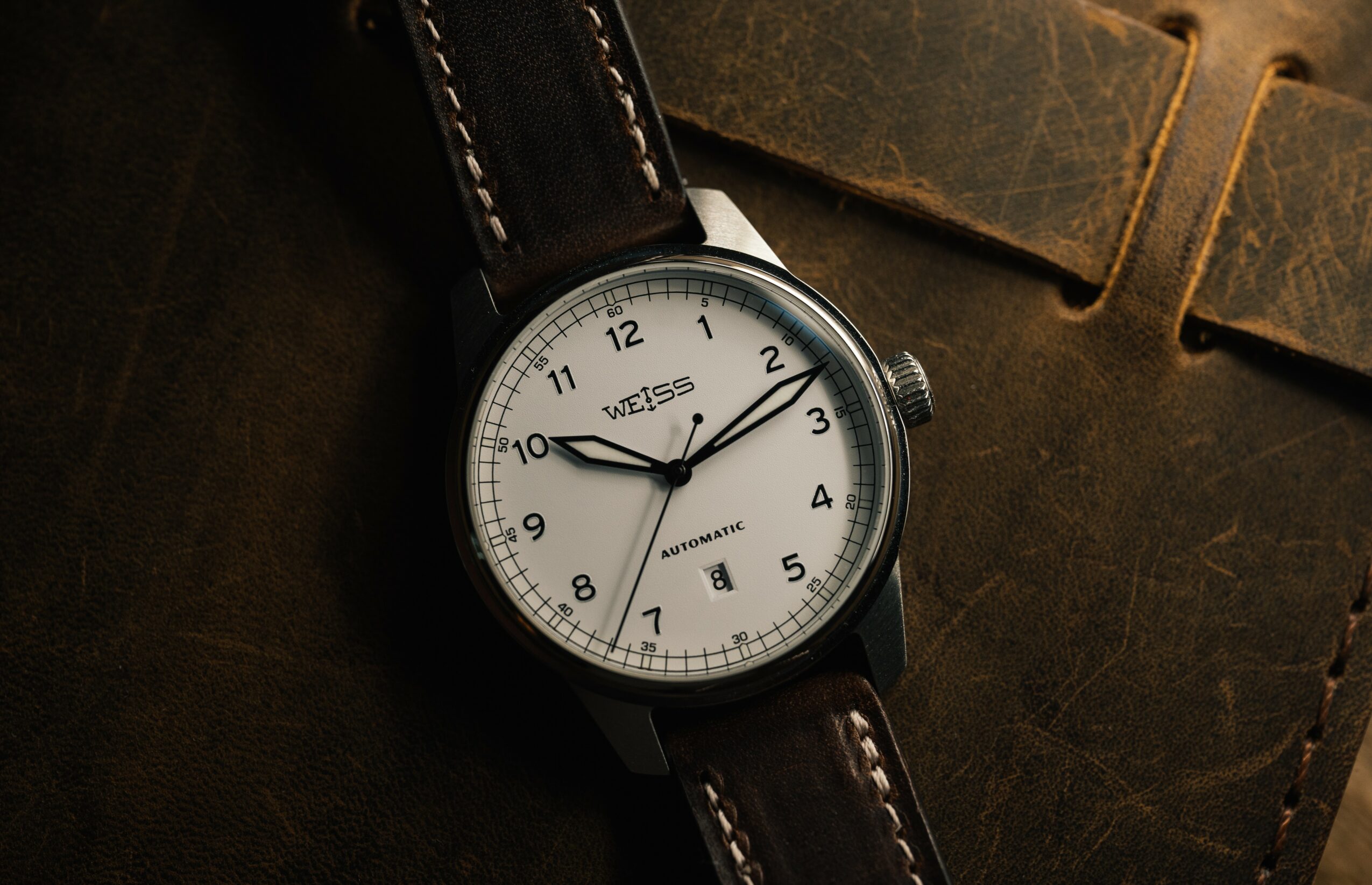 Watch with white face, black case, and brown leather strap on dark background.
