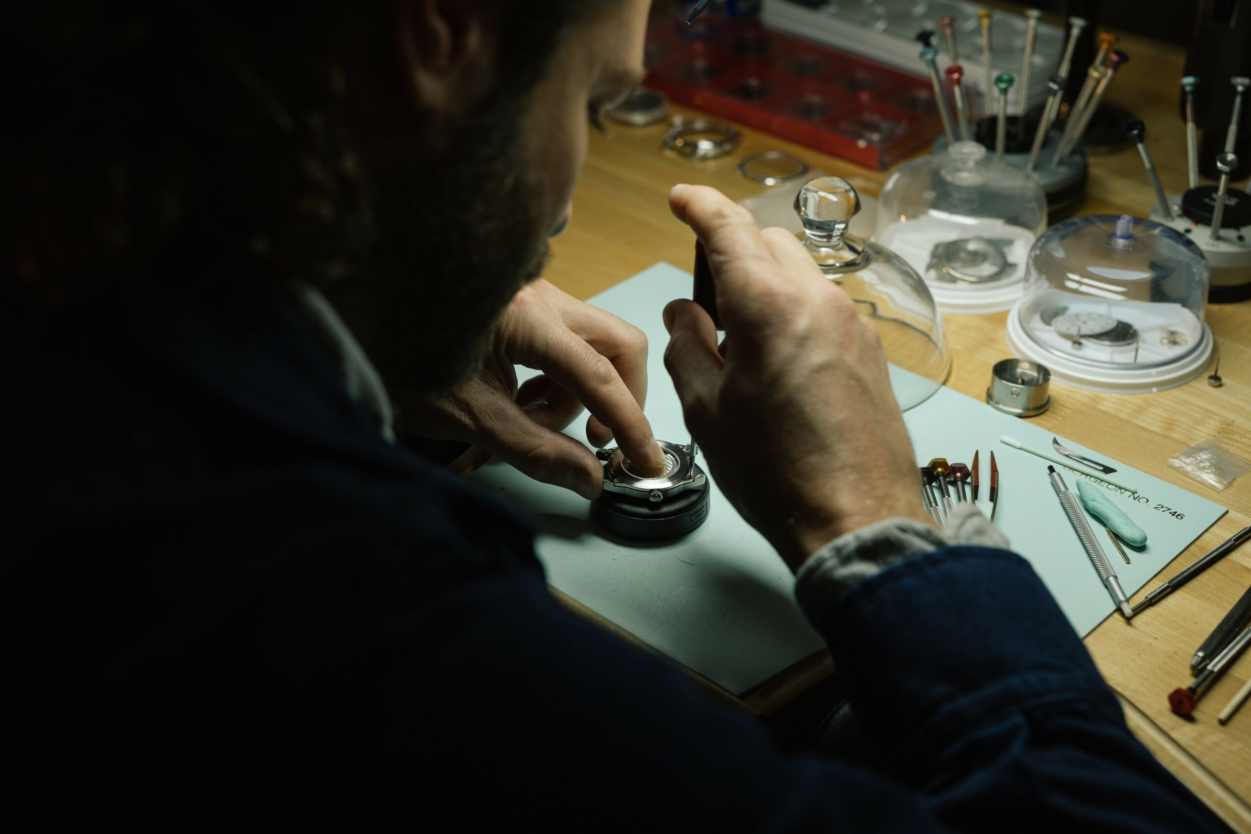 Person working on a watch repair at a cluttered workbench with tools and watch parts.