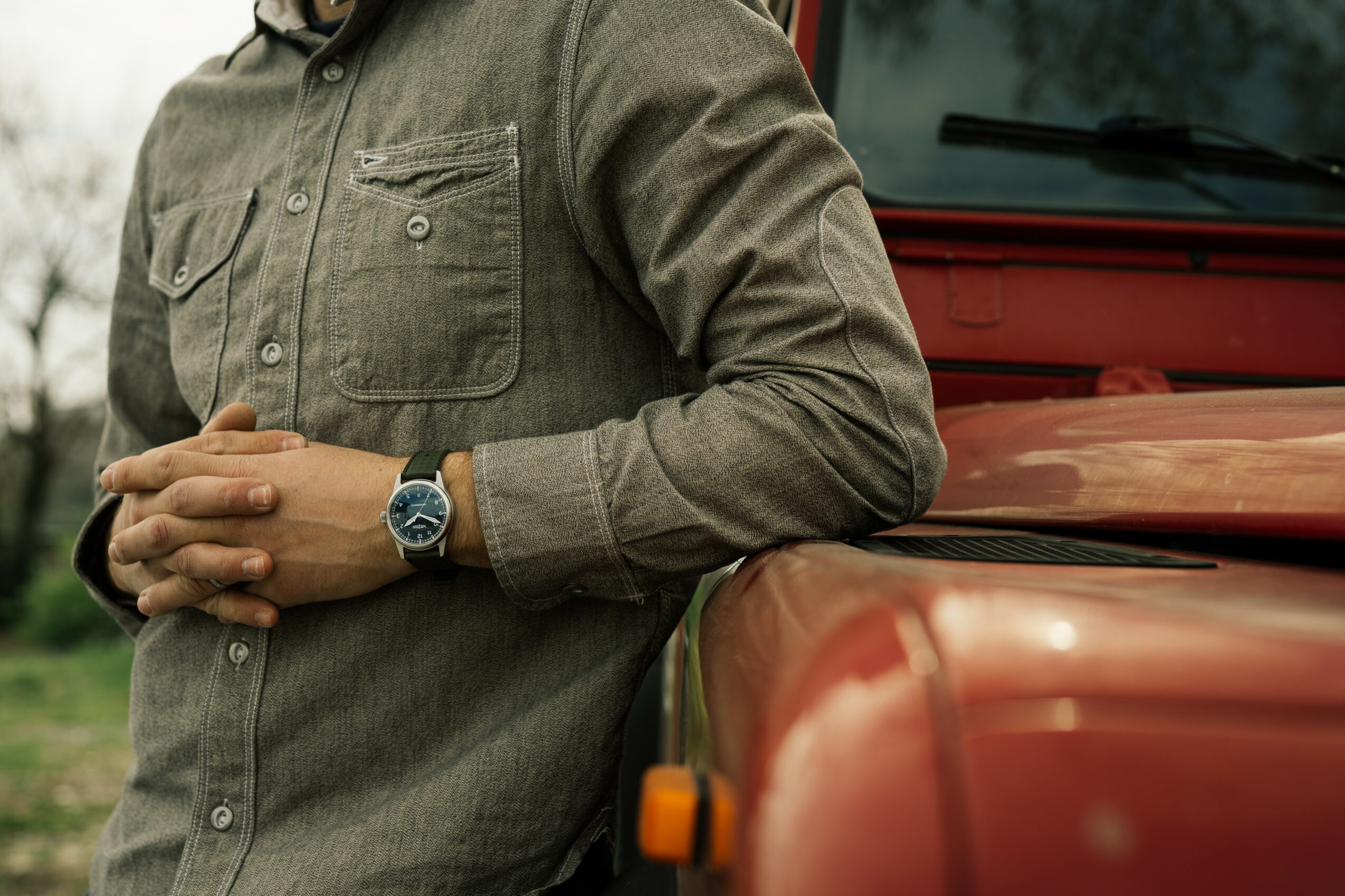 Person wearing a long-sleeve shirt with a watch, leaning against a red vehicle, with hands clasped.