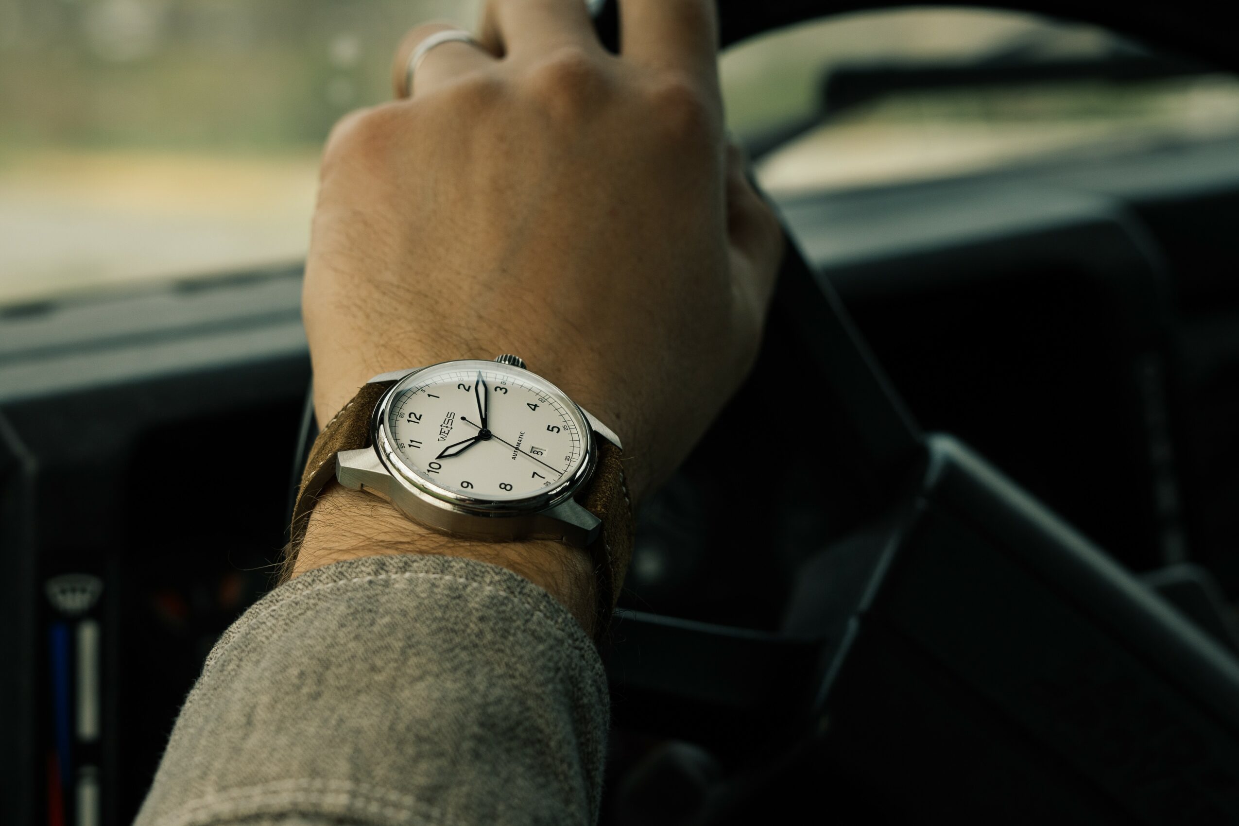 Person's hand on steering wheel with watch on wrist, inside a vehicle, view through windshield, daytime.