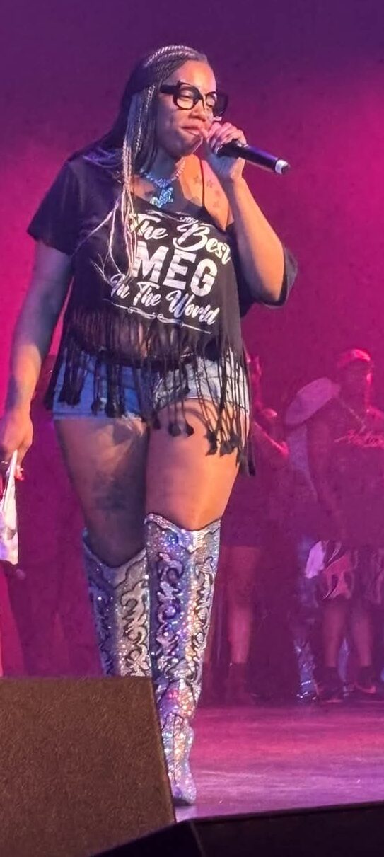 A woman with long hair and glasses sings into a microphone on stage, wearing a black T-shirt and patterned thigh-high boots.