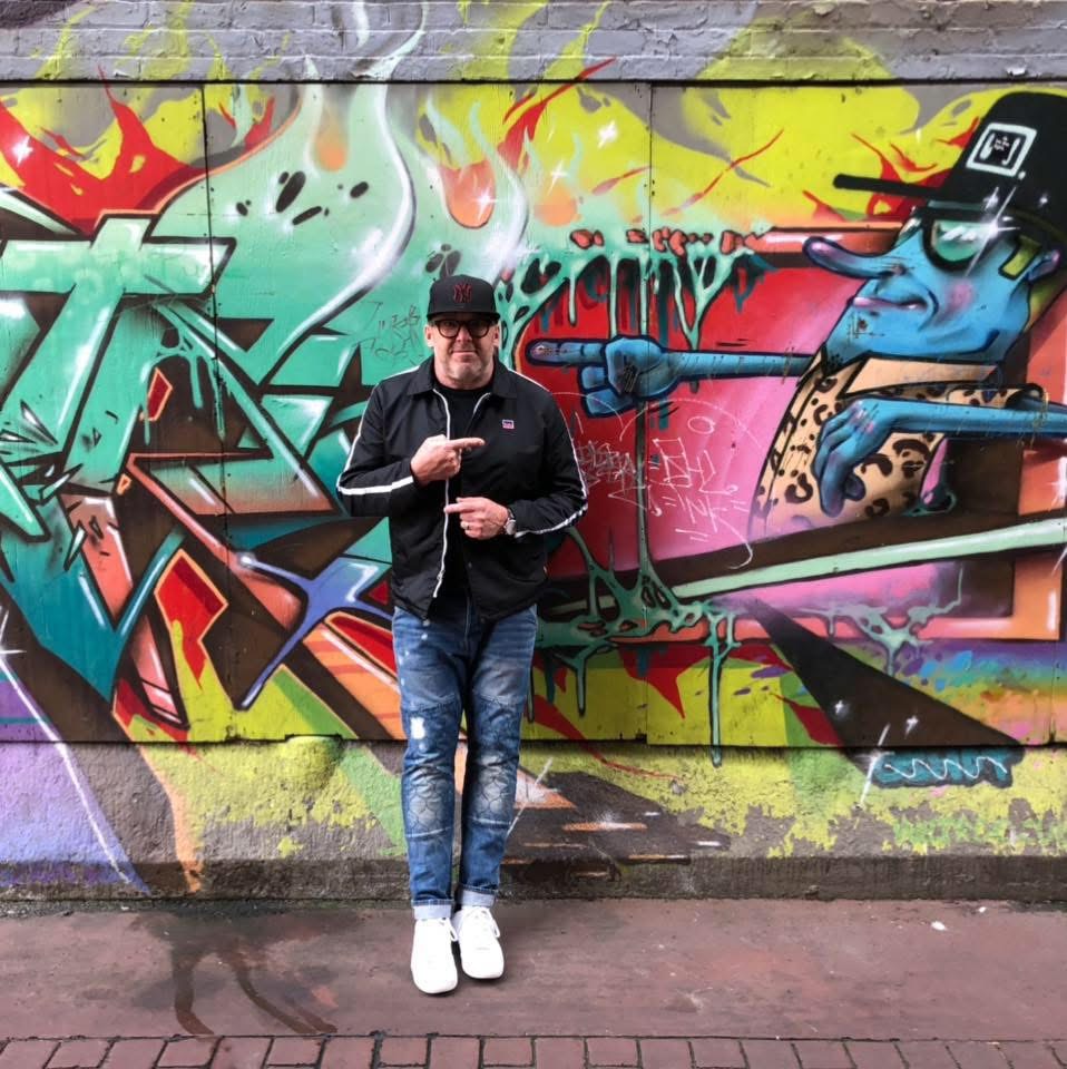 Person standing in front of colorful graffiti wall, wearing black jacket, jeans, white shoes, and cap, holding a phone.