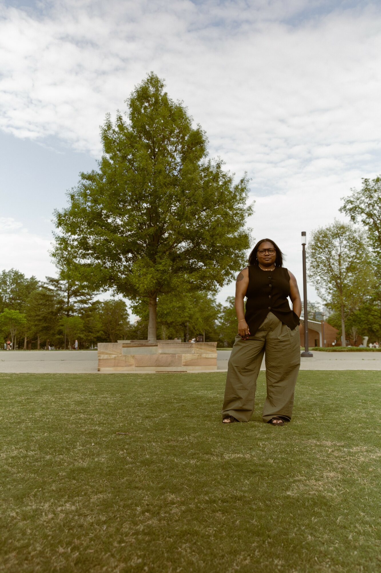 Person standing on grass in front of a large tree with a cloudy sky background.