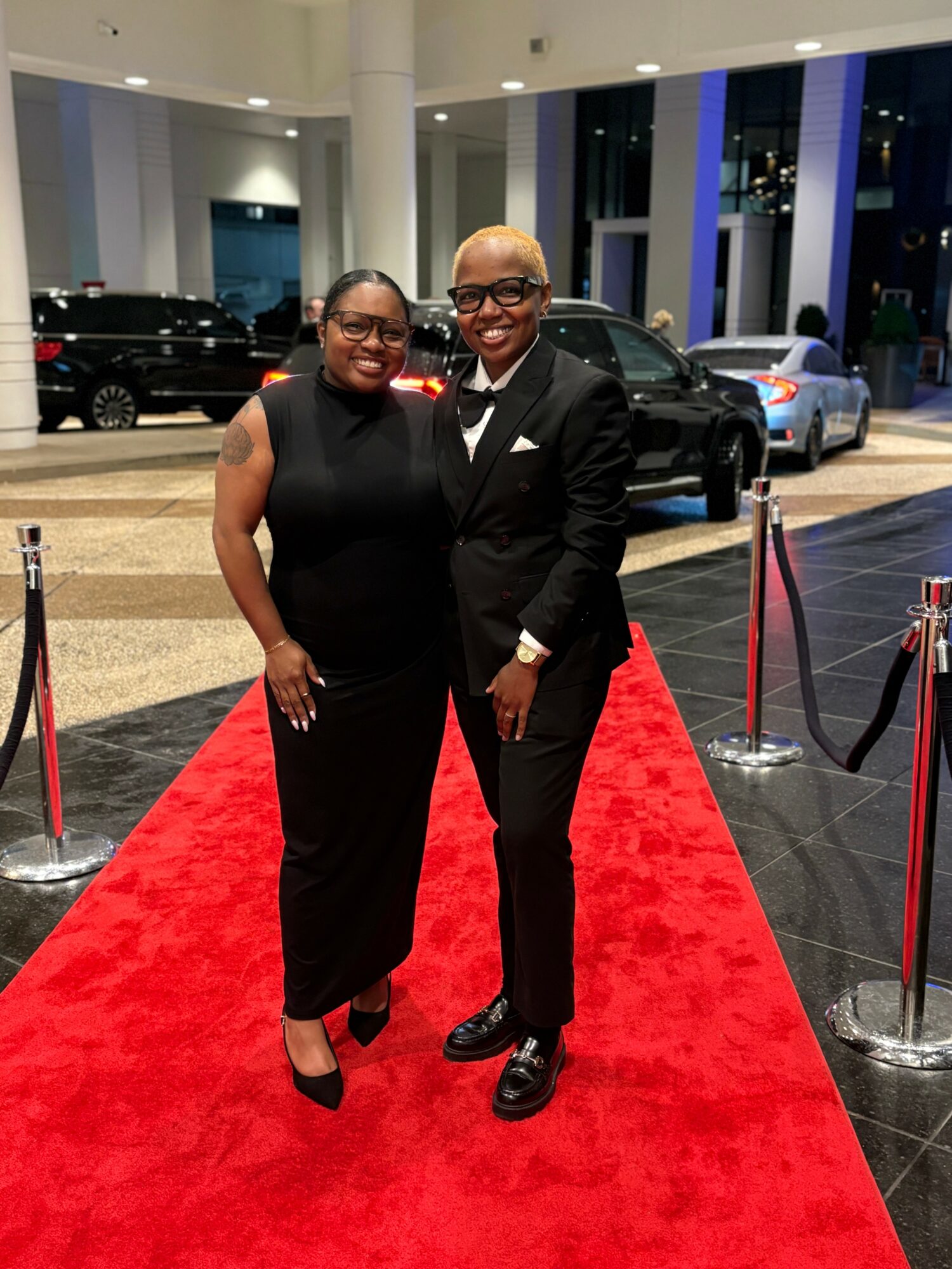 Two people in formal attire standing on a red carpet inside a building with large windows and parked cars outside.
