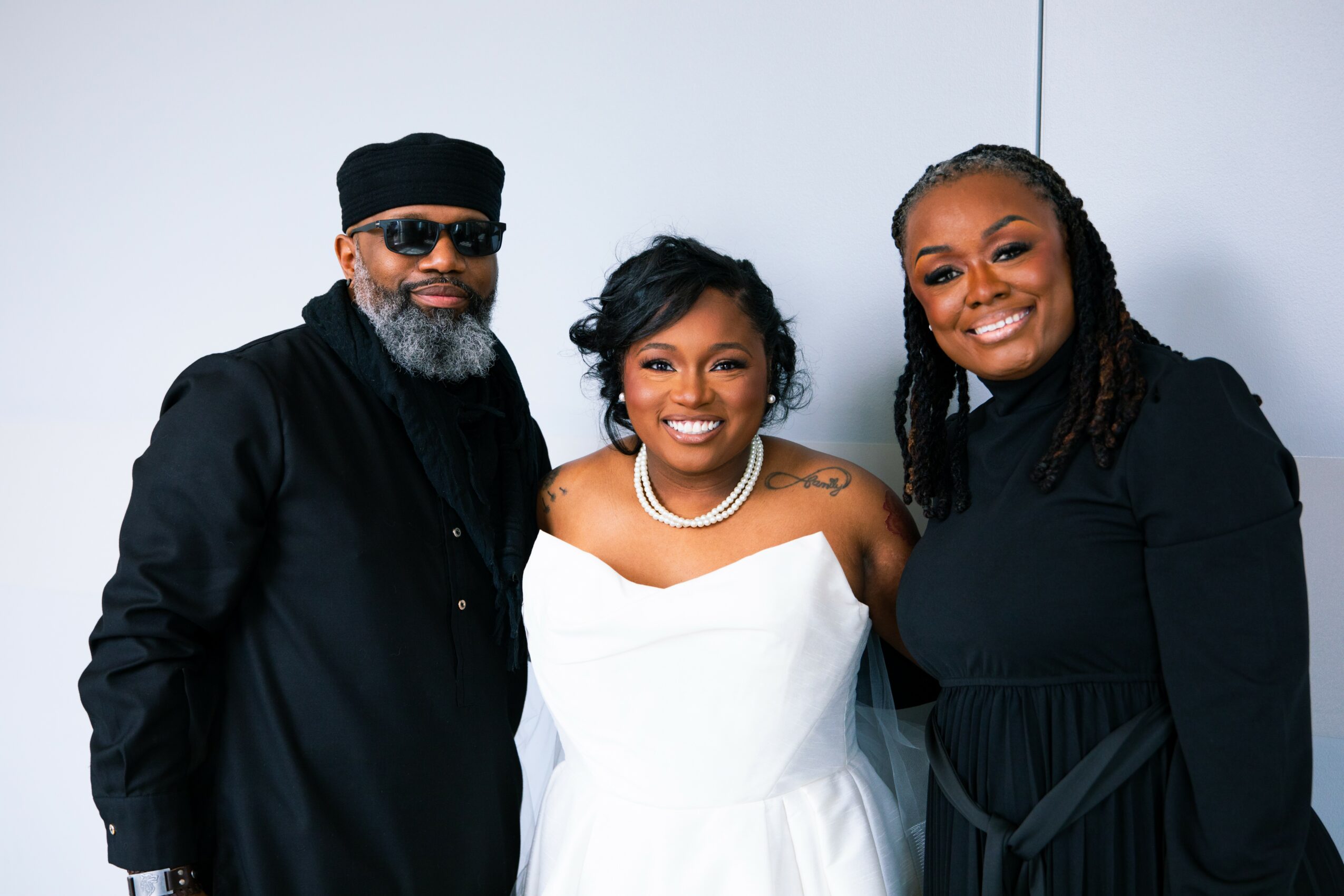 Three people smiling, standing close together against a plain background, dressed in black and white clothing.
