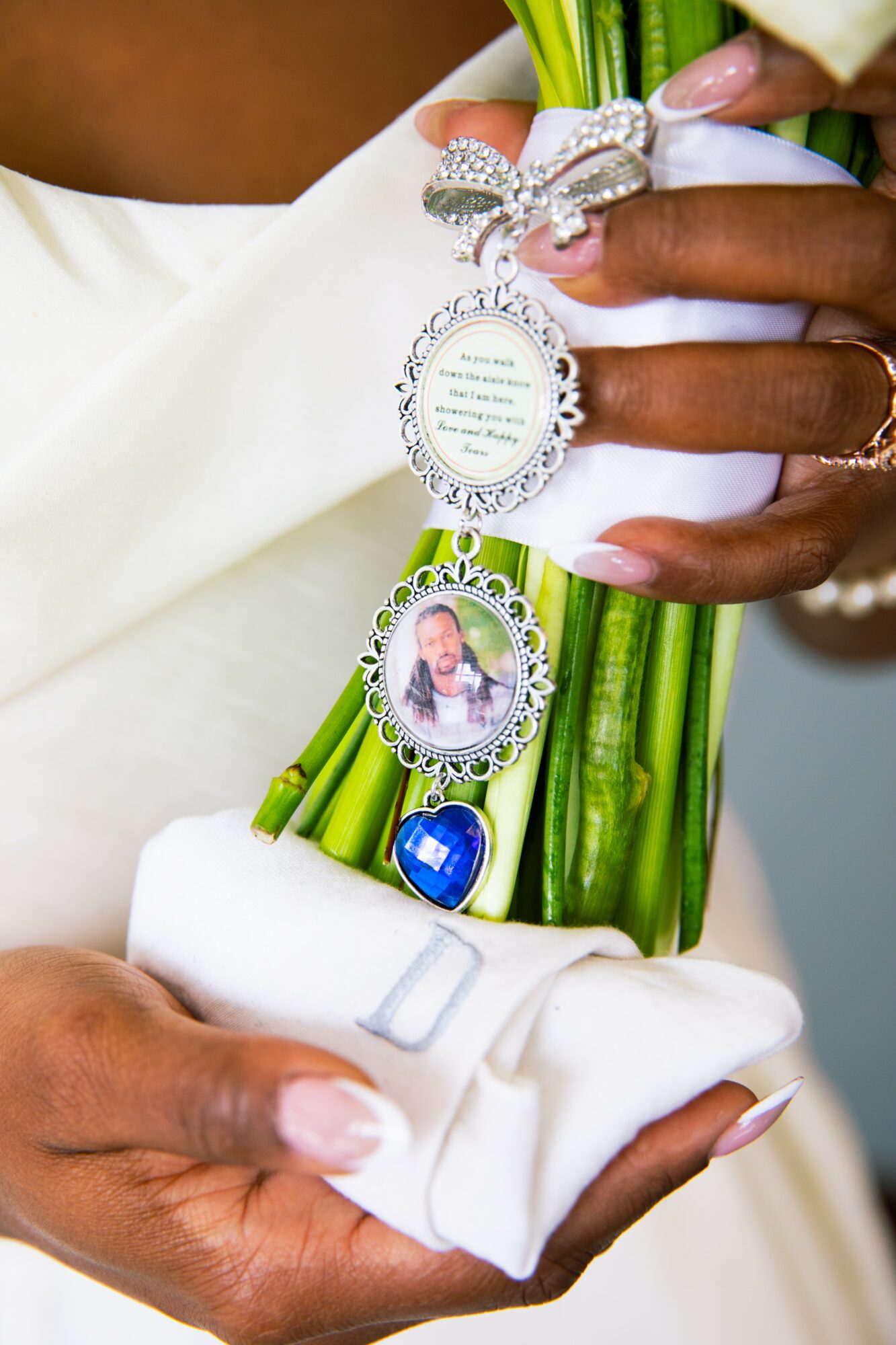 Person holding a bouquet with a religious medal, a photo charm, and a blue heart charm, wearing white gloves.