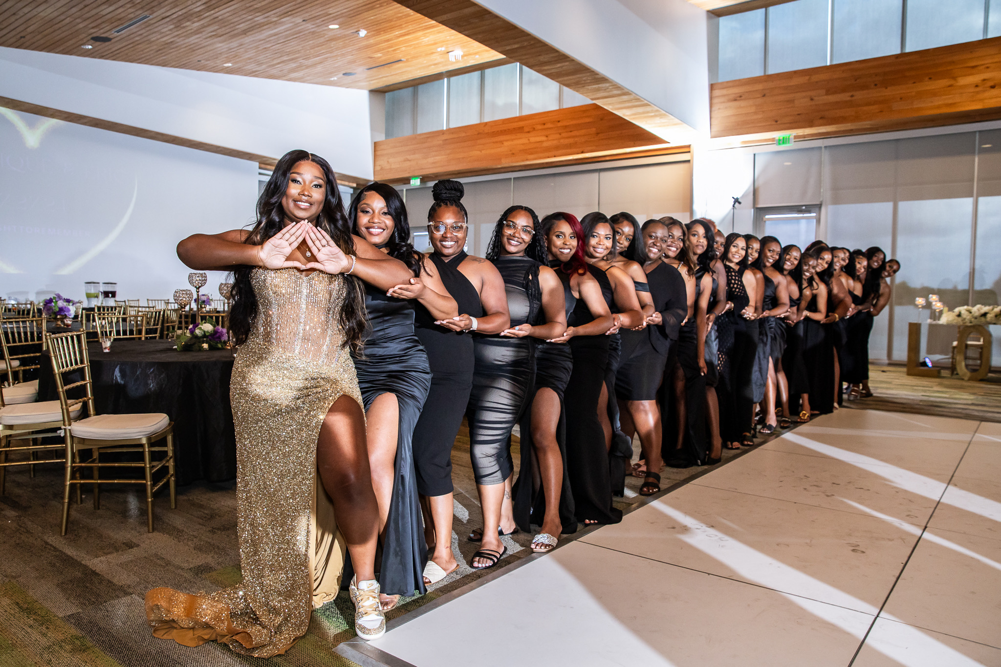 Group of women in formal attire standing in a line indoors, smiling and posing for a photo, with tables and chairs in the background.