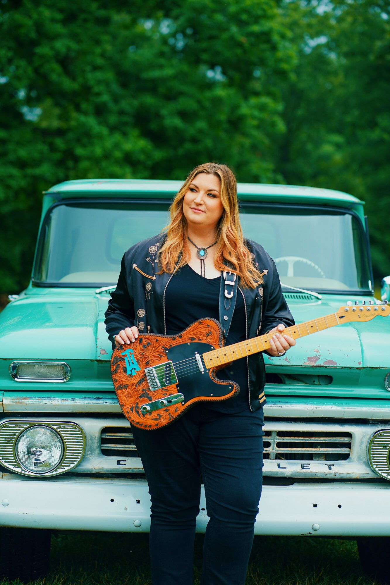 Woman with long hair holding a guitar in front of a vintage vehicle outdoors, with green trees in background.
