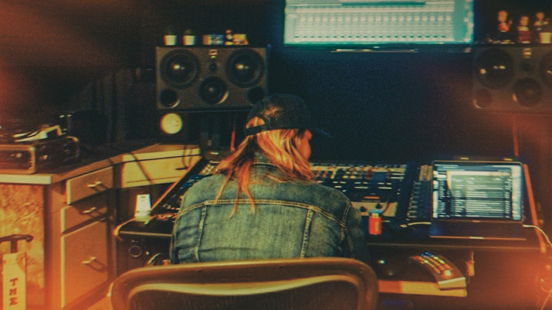 Person with long hair wearing a cap and denim jacket working at a music studio with equipment and screens