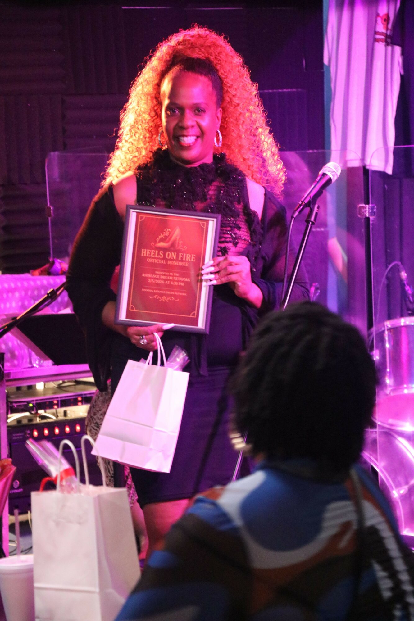 Woman with curly hair holding a framed certificate on stage, with a person in the foreground, pink lighting, and a microphone stand.