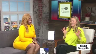 Two women sitting on a TV set, smiling and talking, with a screen behind them displaying a logo and text.