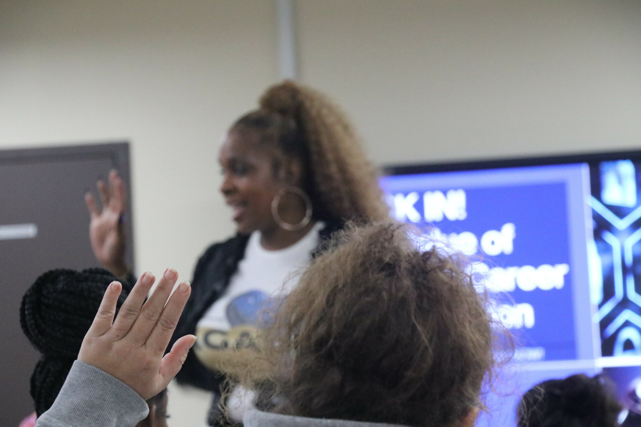 Two women are in a room, one with curly hair and large earrings, the other with braided hair, raising their hands.