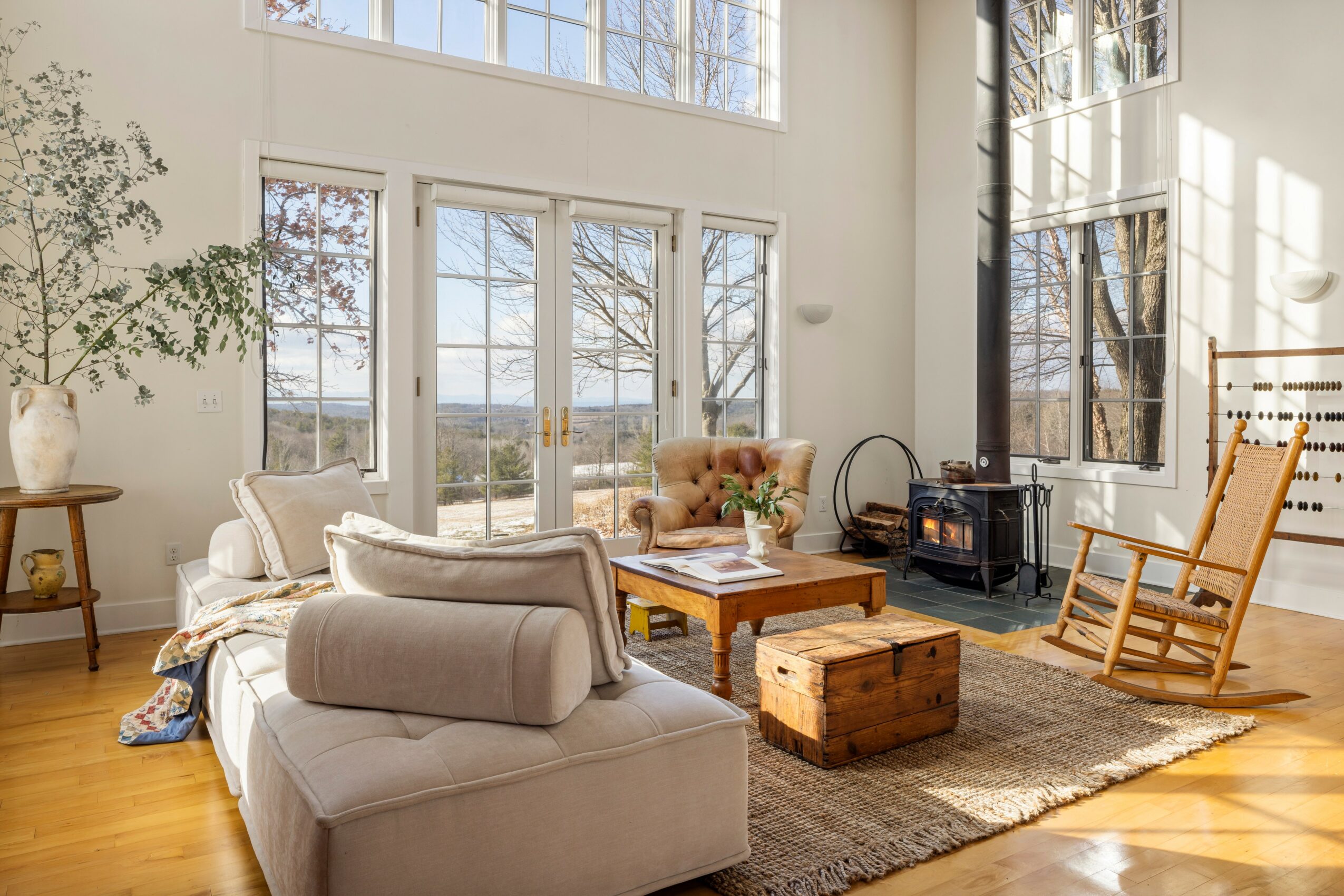 Bright living room with large windows, beige sofa, wooden rocking chair, and a wooden chest, sunlight streaming in.