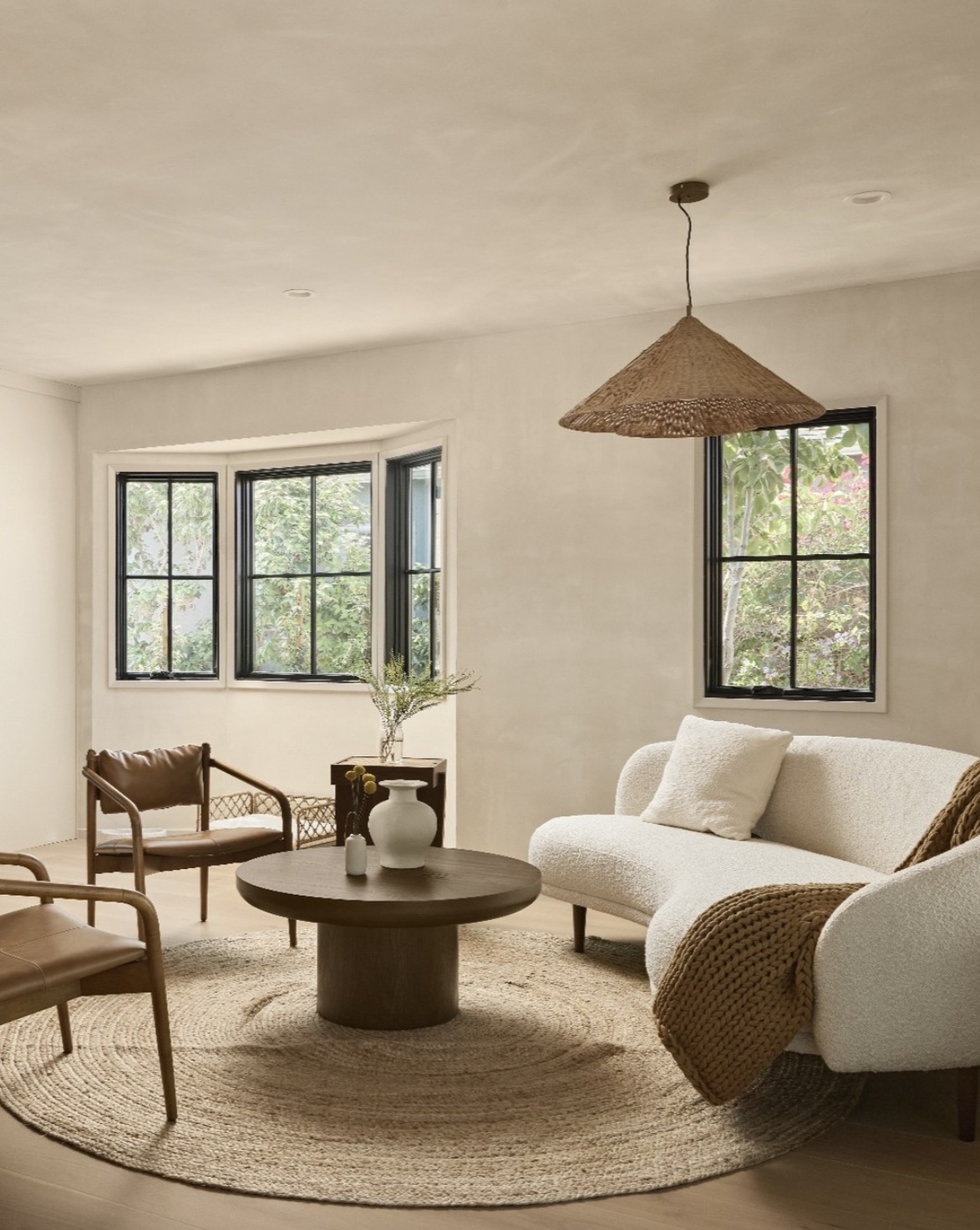 Living room with large windows, white walls, a round rug, a white sofa, wooden chairs, and a hanging light fixture.