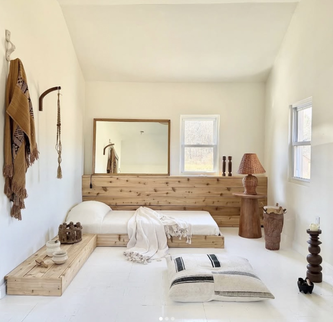 Minimalist bedroom with a low bed, wooden headboard, mirror, window, and decorative items, in neutral tones.