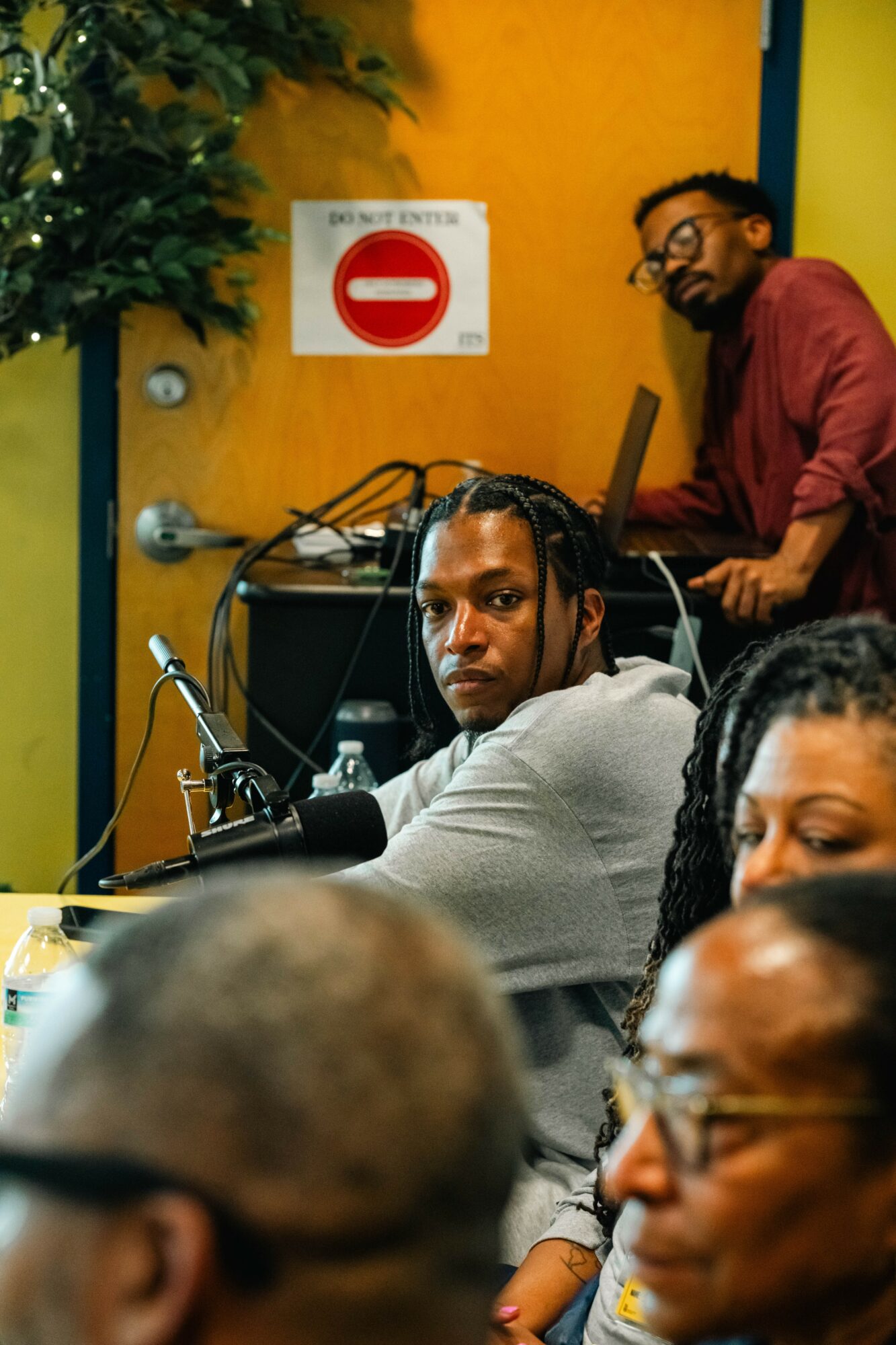 Group of people in a room with a yellow wall, a 'no entry' sign, and a man with glasses working on a laptop.