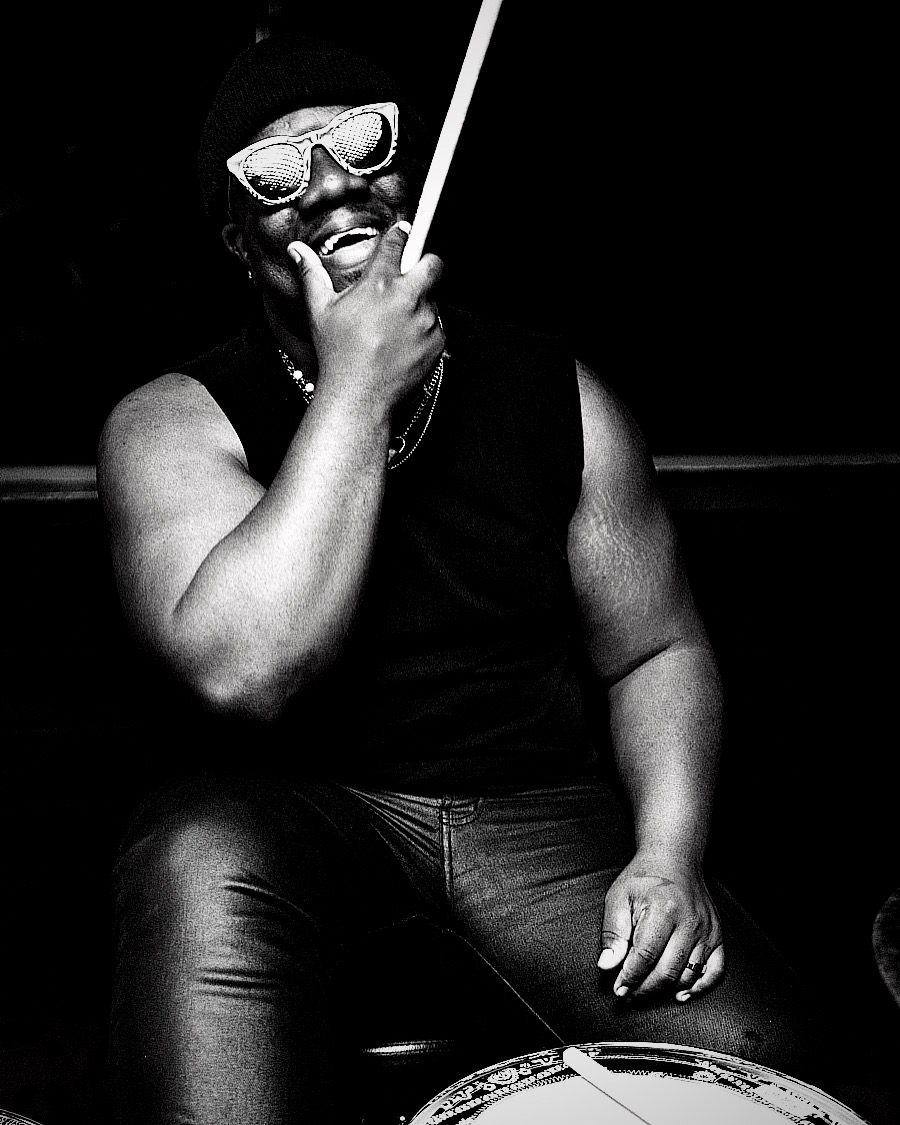 Person wearing sunglasses and a sleeveless shirt, holding a stick near their face, sitting in a dark environment.