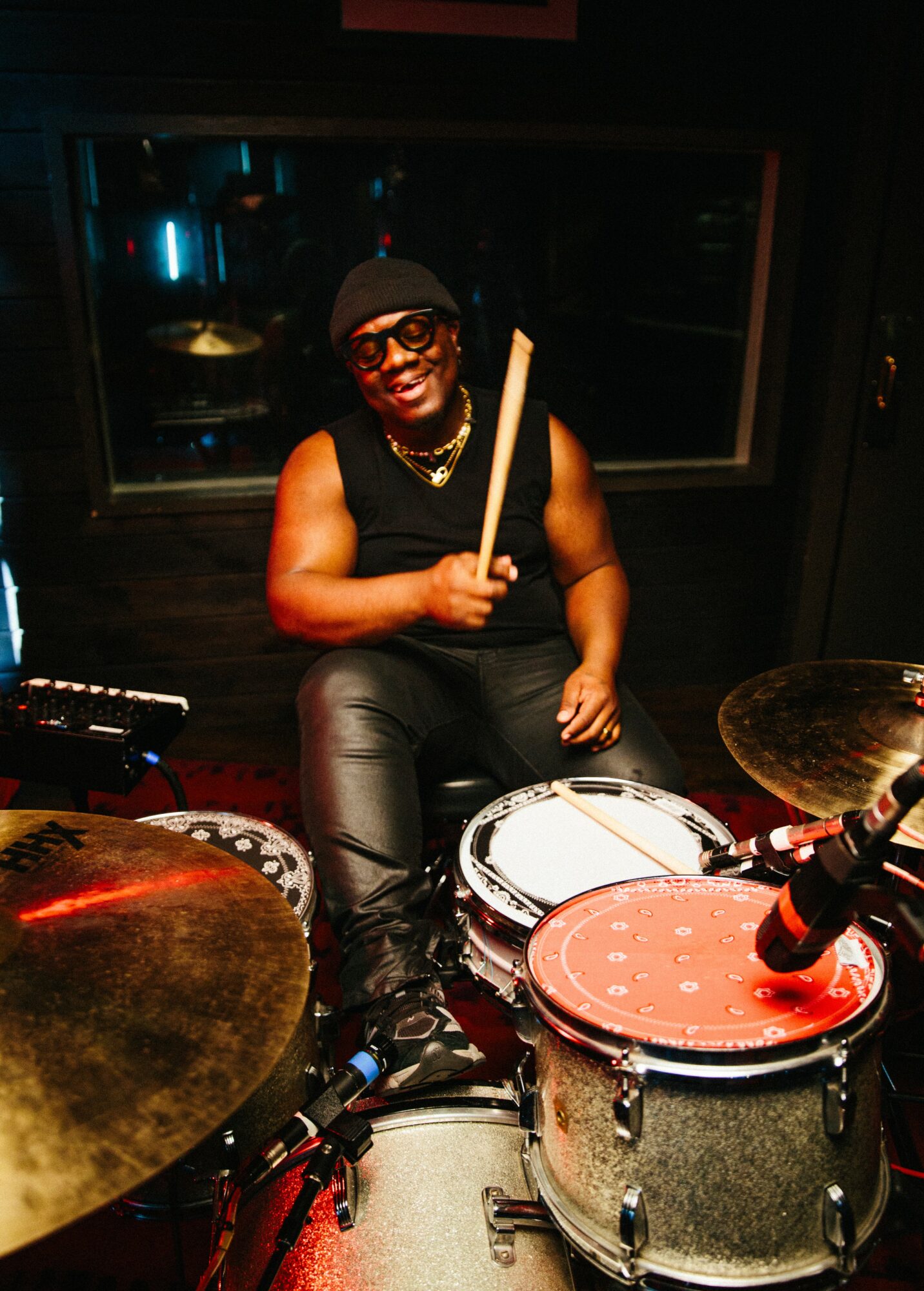 Person sitting behind a drum set, holding a drumstick, smiling, wearing glasses and a sleeveless top, in a dark room.