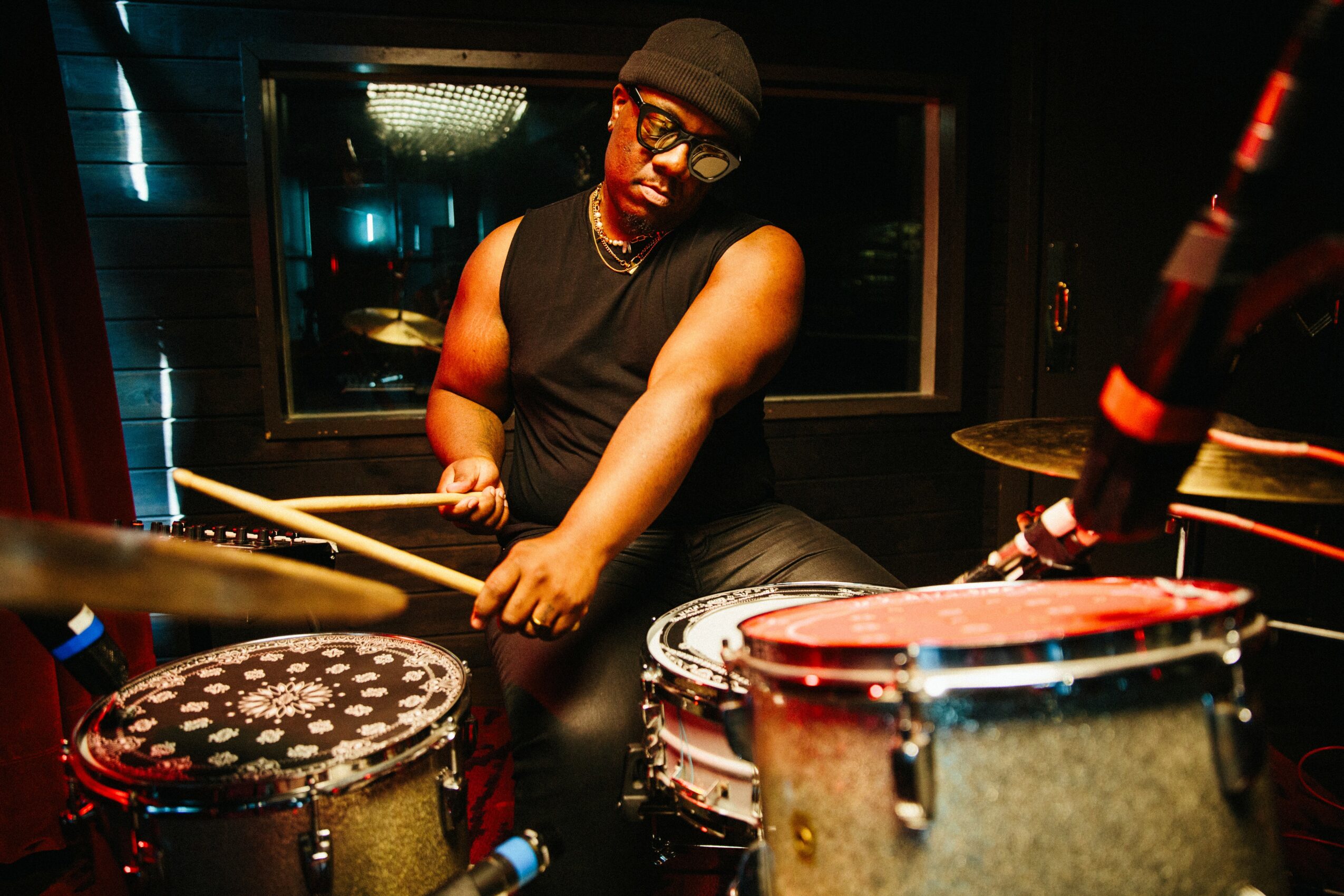 Person playing drums in a recording studio, wearing a black sleeveless shirt and glasses, with a focused expression.