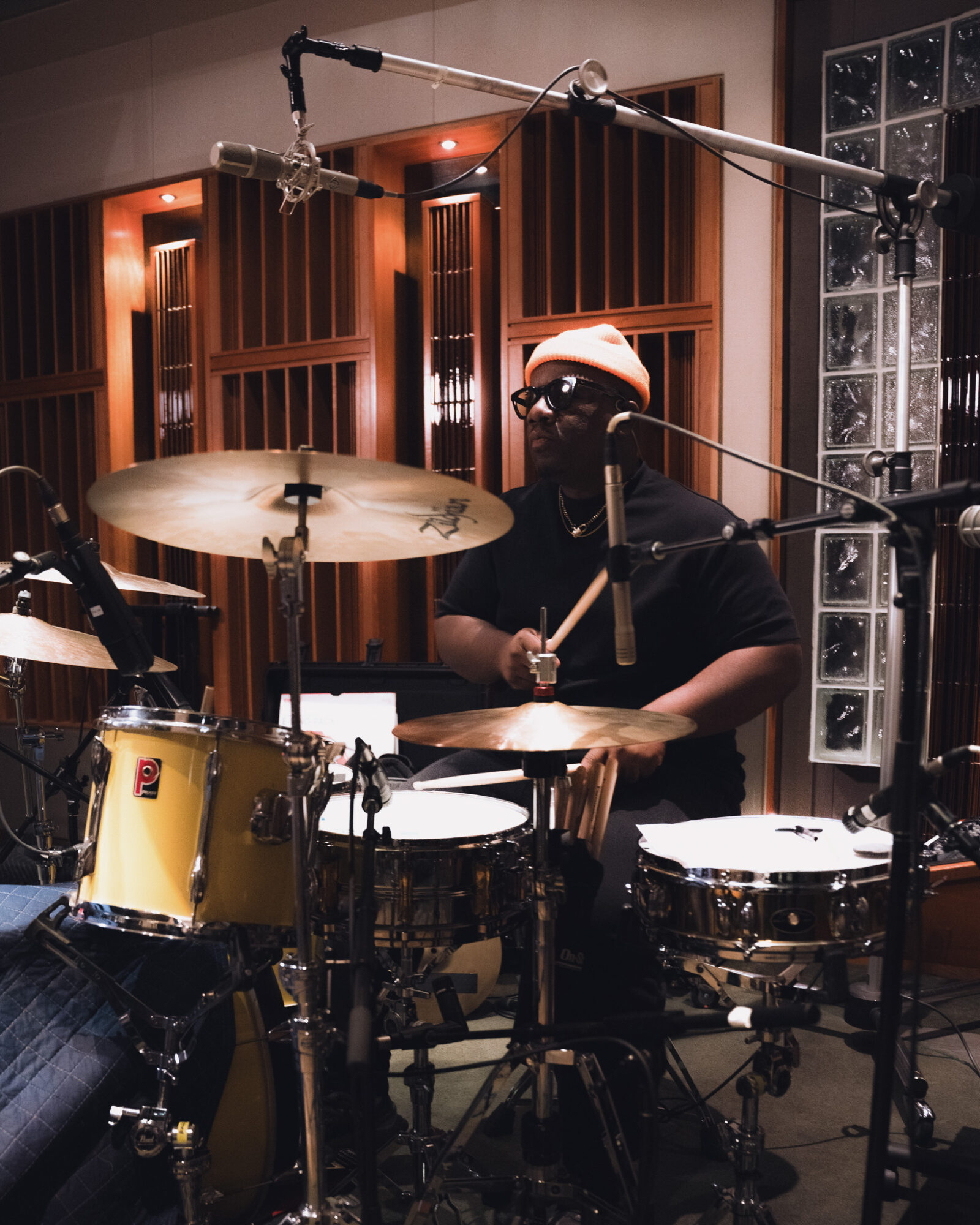 Person playing drums in a recording studio with wooden panels and glass blocks, wearing a hat and glasses.