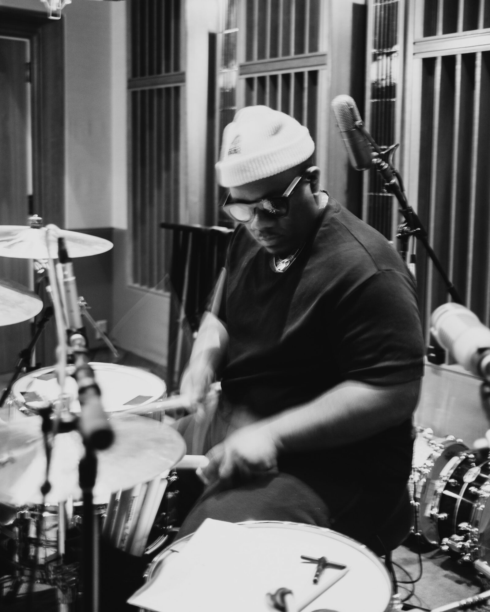 Person playing drums in a recording studio, wearing a beanie and glasses, surrounded by musical equipment.