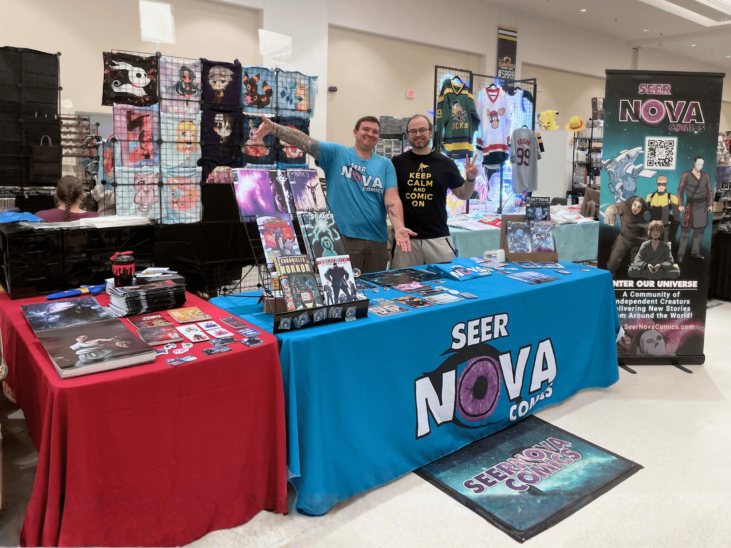 Two people stand behind a table with merchandise at a convention, with a Seer Nova banner and display in the background.