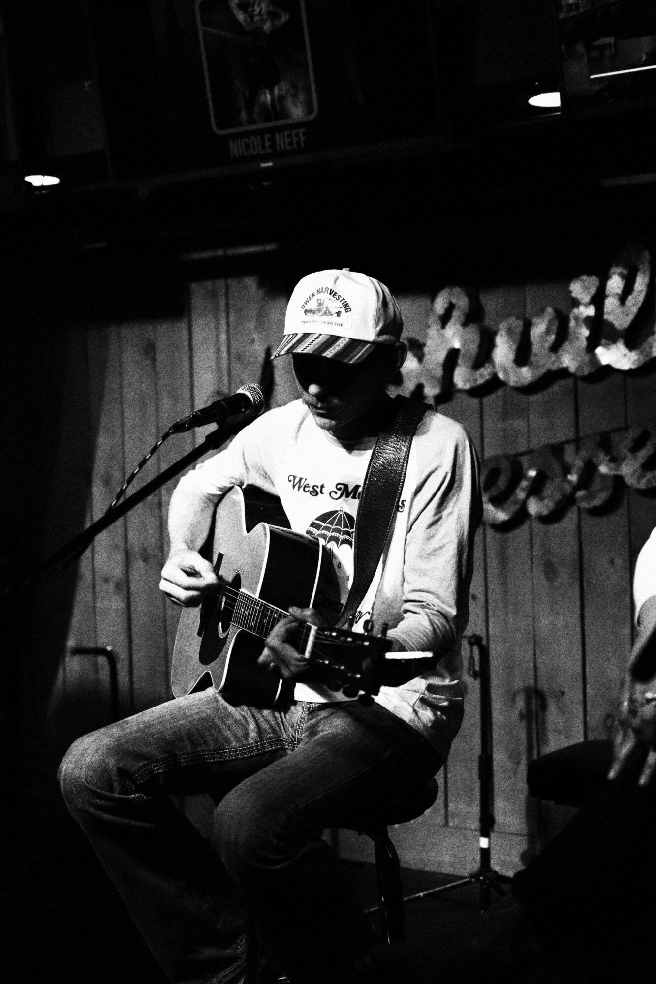 Person sitting on a wooden floor playing an acoustic guitar, wearing a cap and casual clothing, with a dark background.