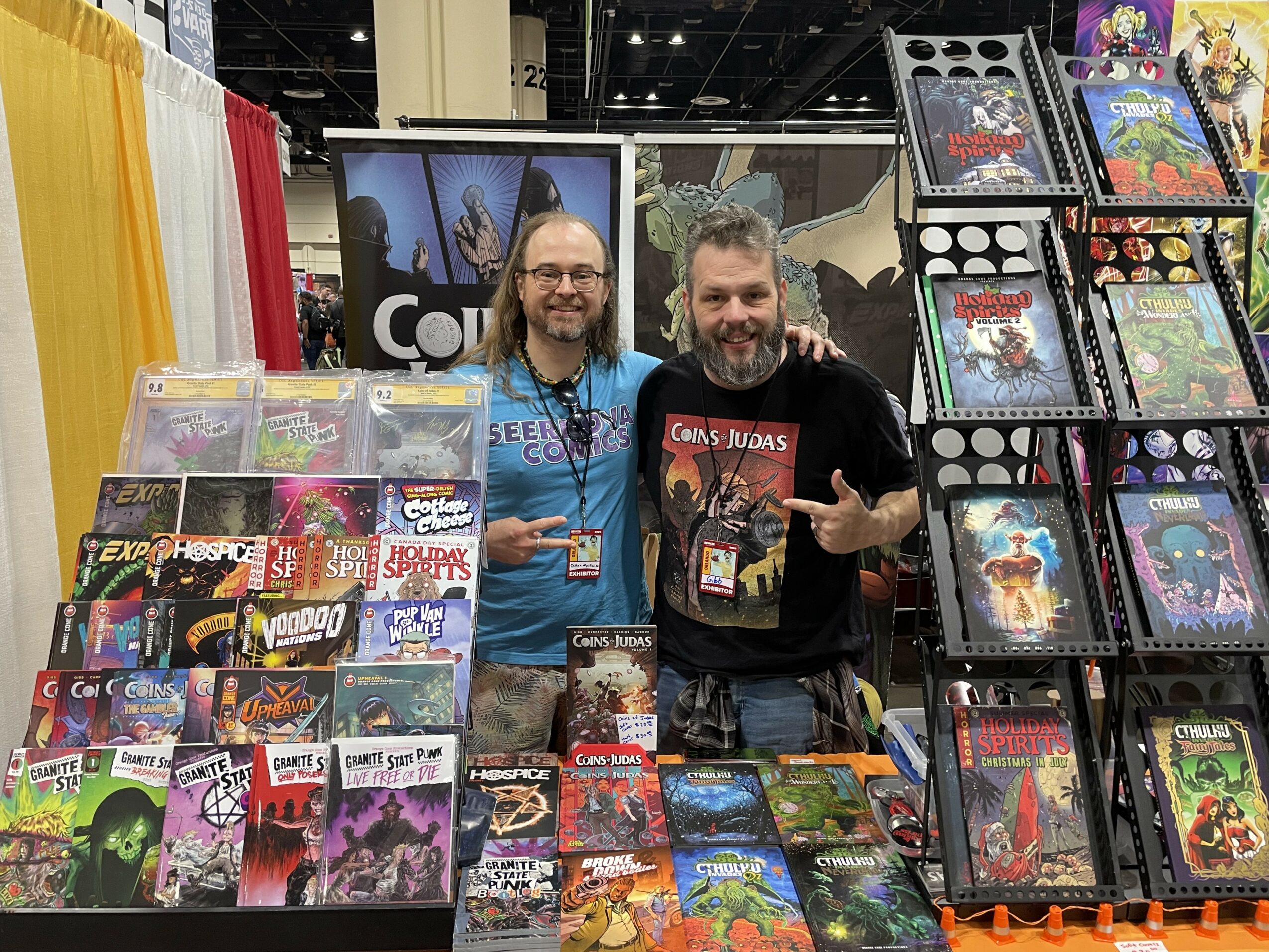 Two men stand behind a table filled with comic books at a convention booth, smiling and posing for the photo.