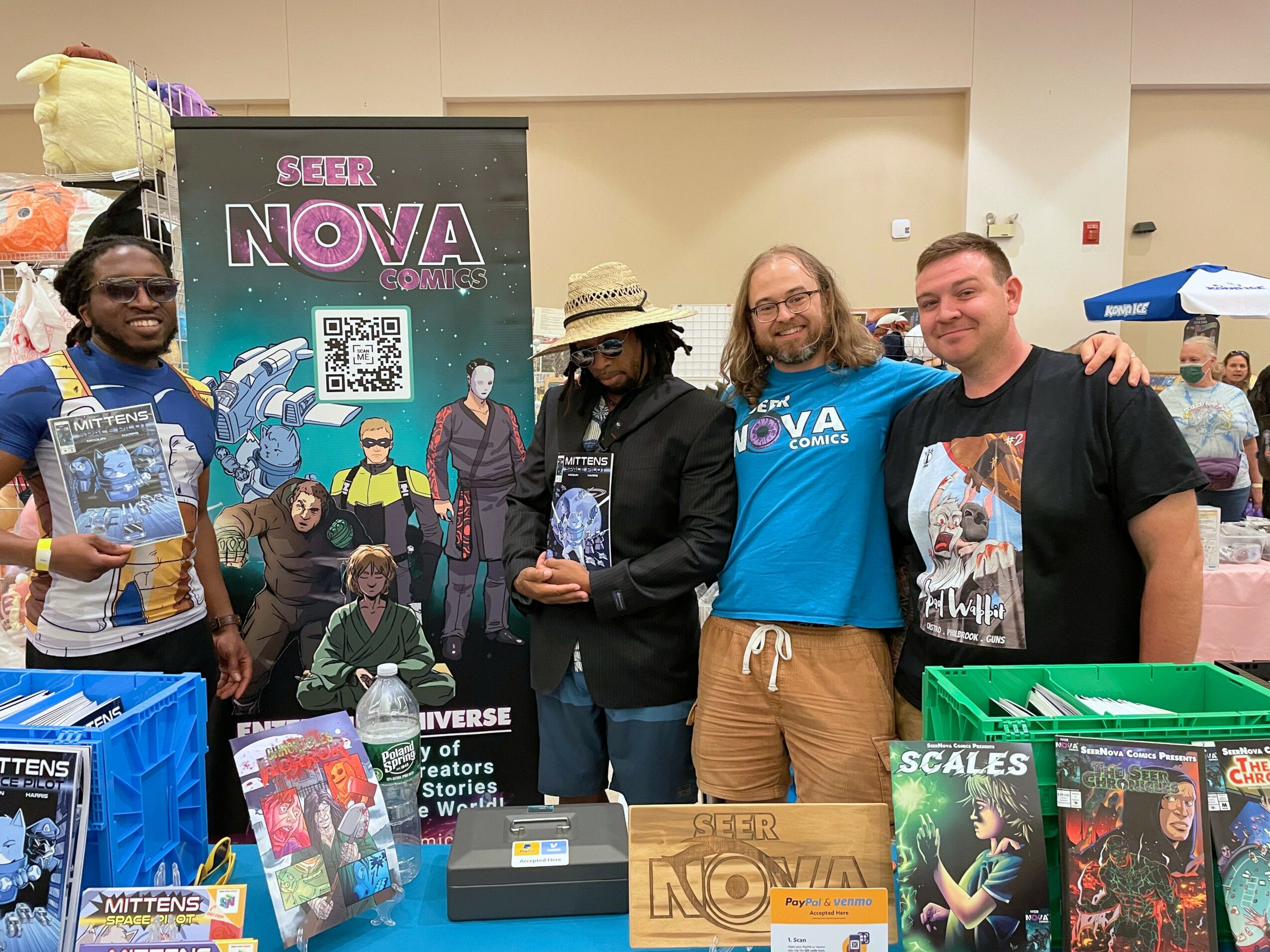 Four people standing indoors, holding comic books, with a SEER NOVA banner behind them. They are smiling and posing for the photo.