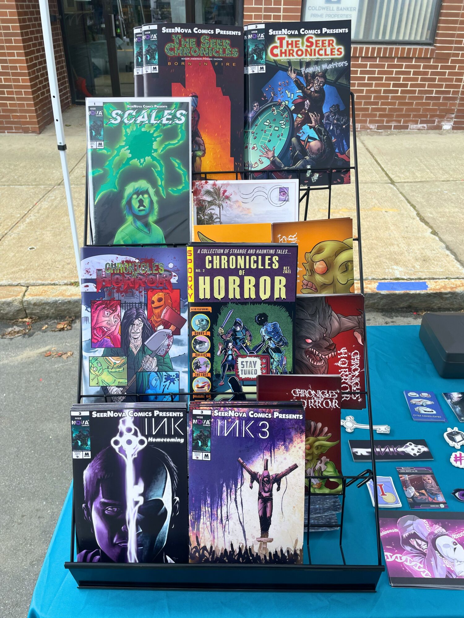 Book display with graphic novels and comics on a metal rack outside, with a brick building in the background.