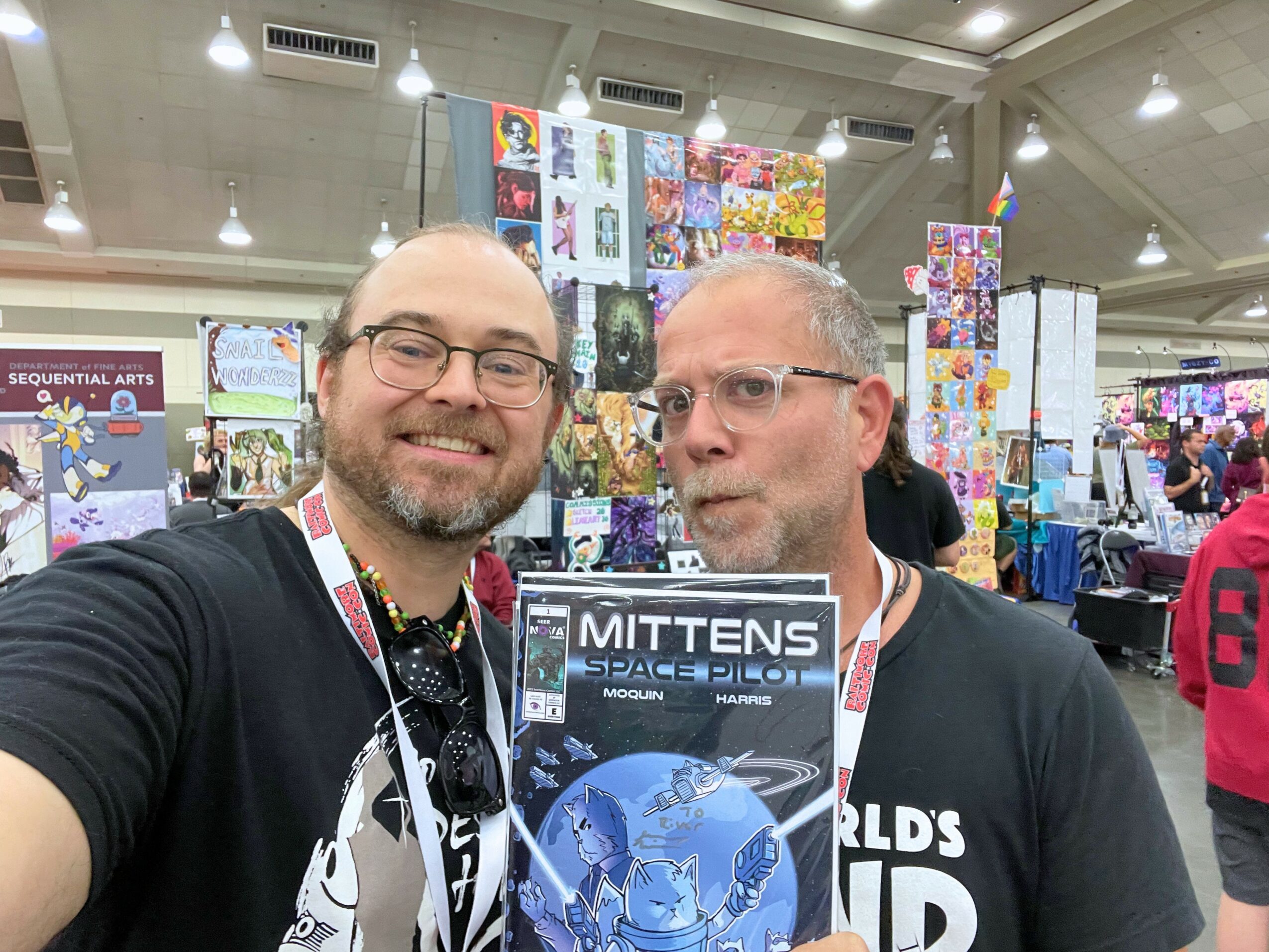 Two men taking a selfie at an indoor event with colorful displays behind them.