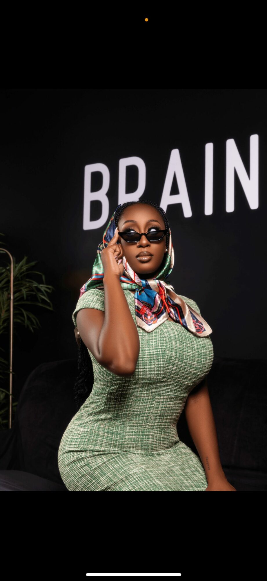 Woman with sunglasses and colorful scarf, standing in front of a black background with white text reading 'BRAIN'.