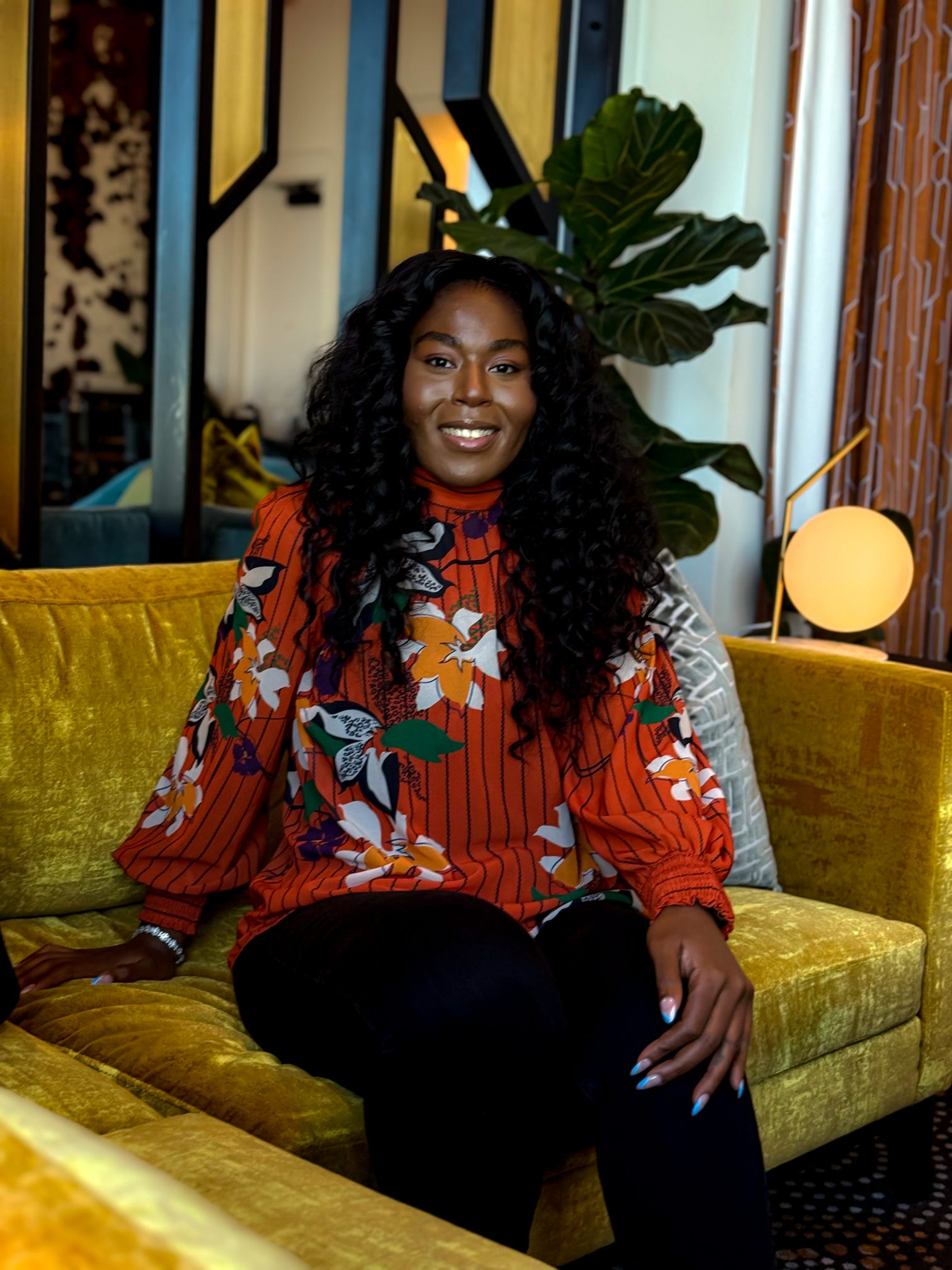 Woman with curly hair sitting on a yellow sofa, smiling, in a room with plants and decorative lighting.