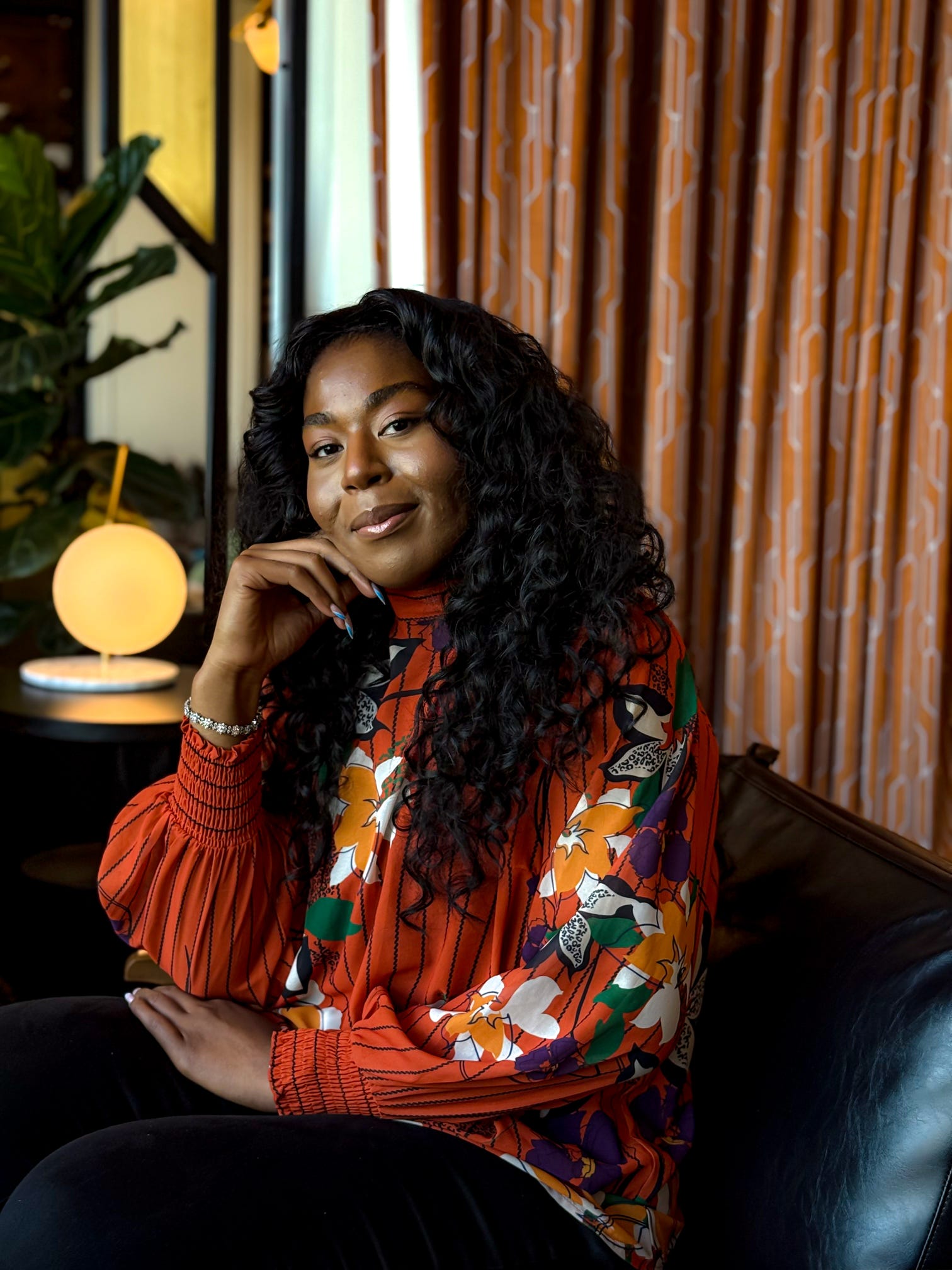 Woman with curly hair sitting on a black chair, wearing a colorful patterned blouse, in a room with curtains and a lamp.