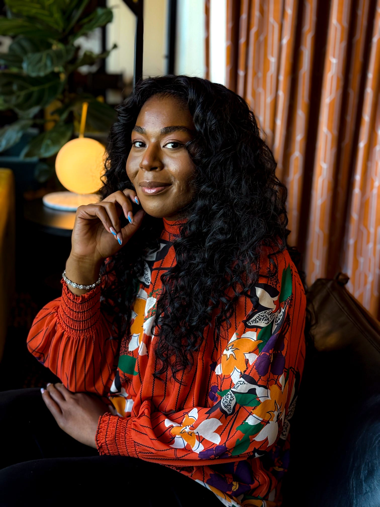 Young woman with long curly hair sitting indoors, wearing a colorful patterned blouse, smiling, with a lamp and curtains in the background.